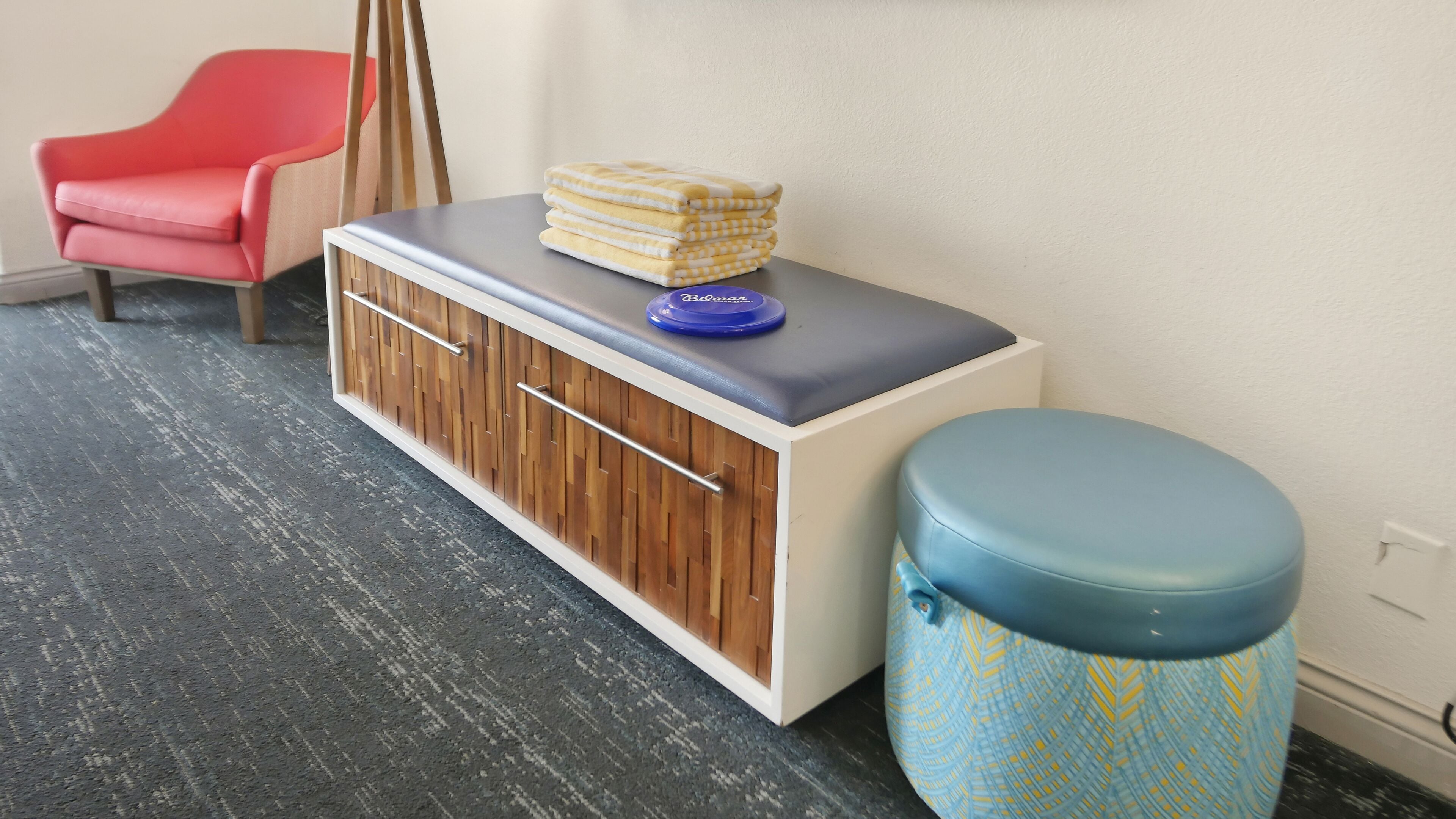 Ein Zimmer mit einem Holzkabinett, einem roten Sessel, einem blauen Hocker und einem Stapel gelb-weiß karierter Handtücher.