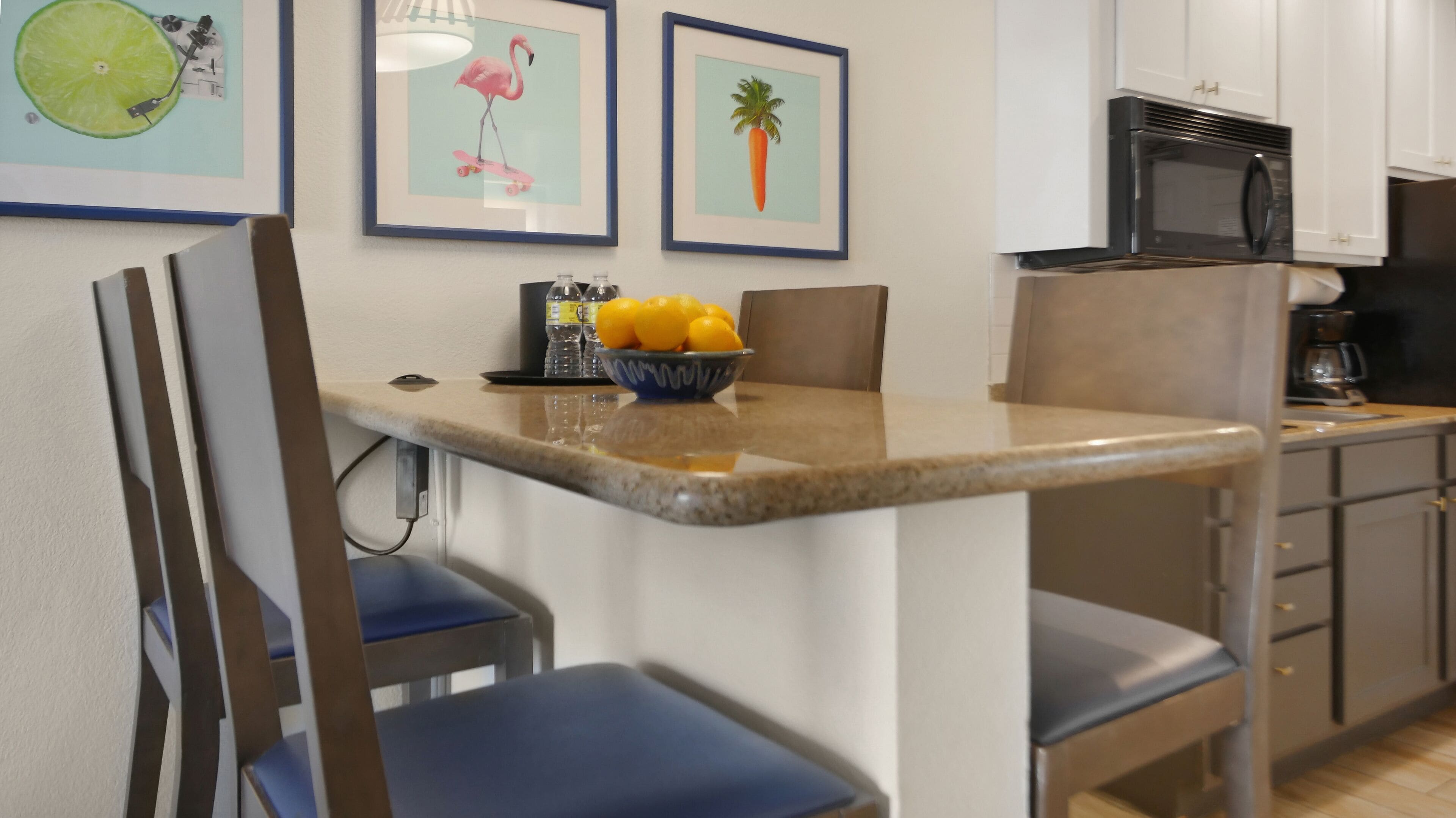 A kitchen with a granite countertop, a bowl of oranges, and a microwave. There are framed pictures on the wall, including a flamingo and a carrot.