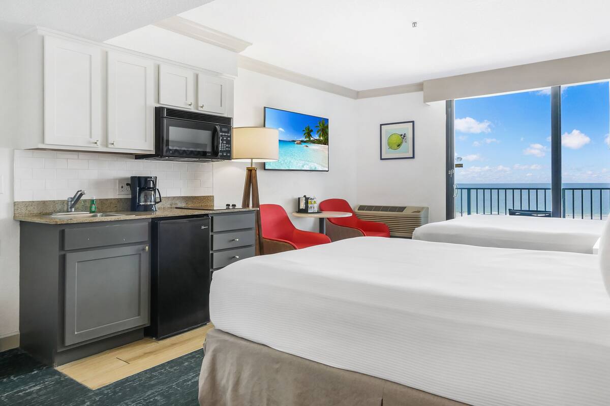 Quarto de hotel com cozinha compacta, duas camas, área de refeições com cadeiras vermelhas e varanda com vista para o mar.