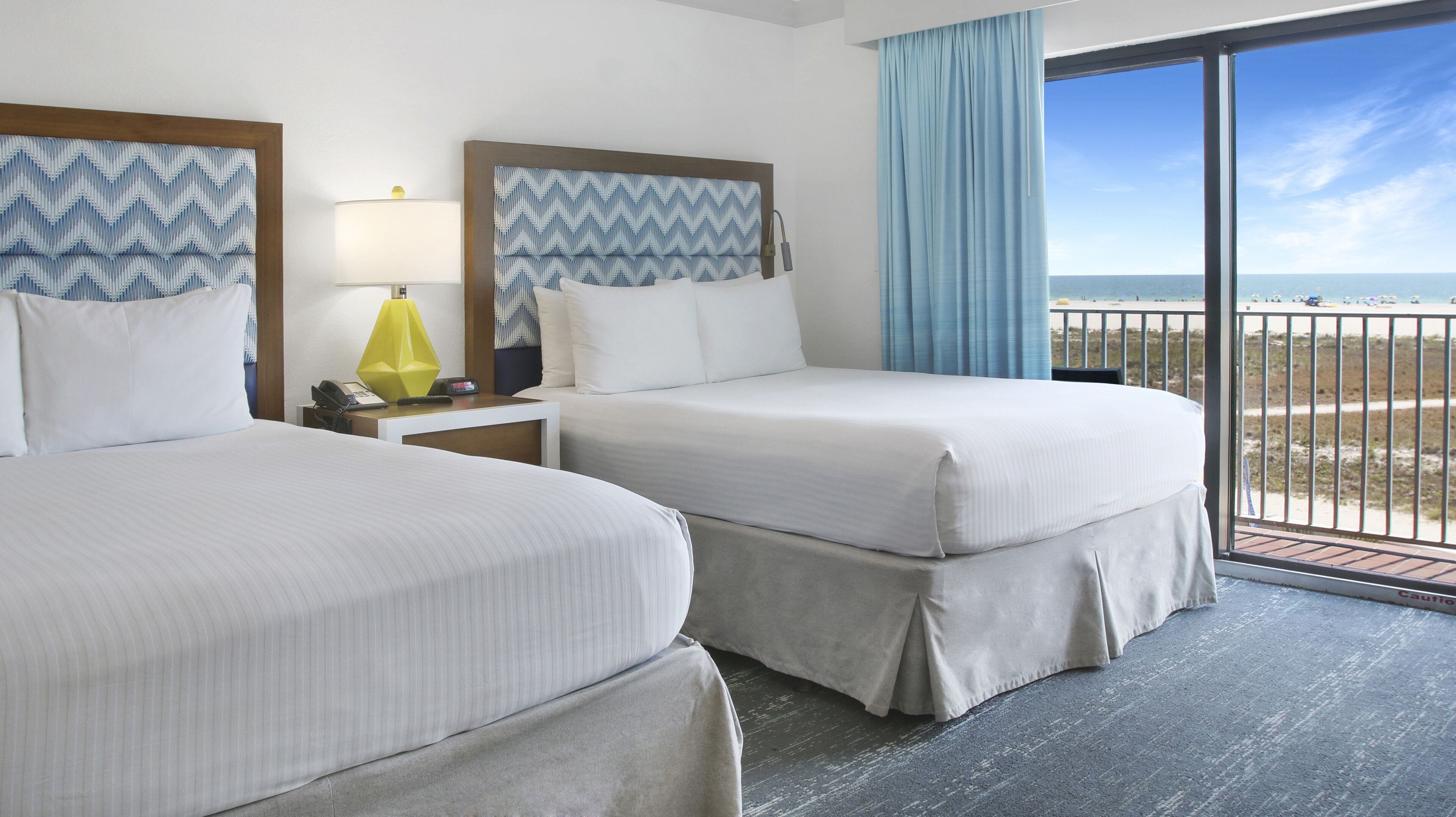 Une chambre d’hôtel avec deux lits, une table de chevet avec une lampe et une vue sur la plage depuis un balcon.