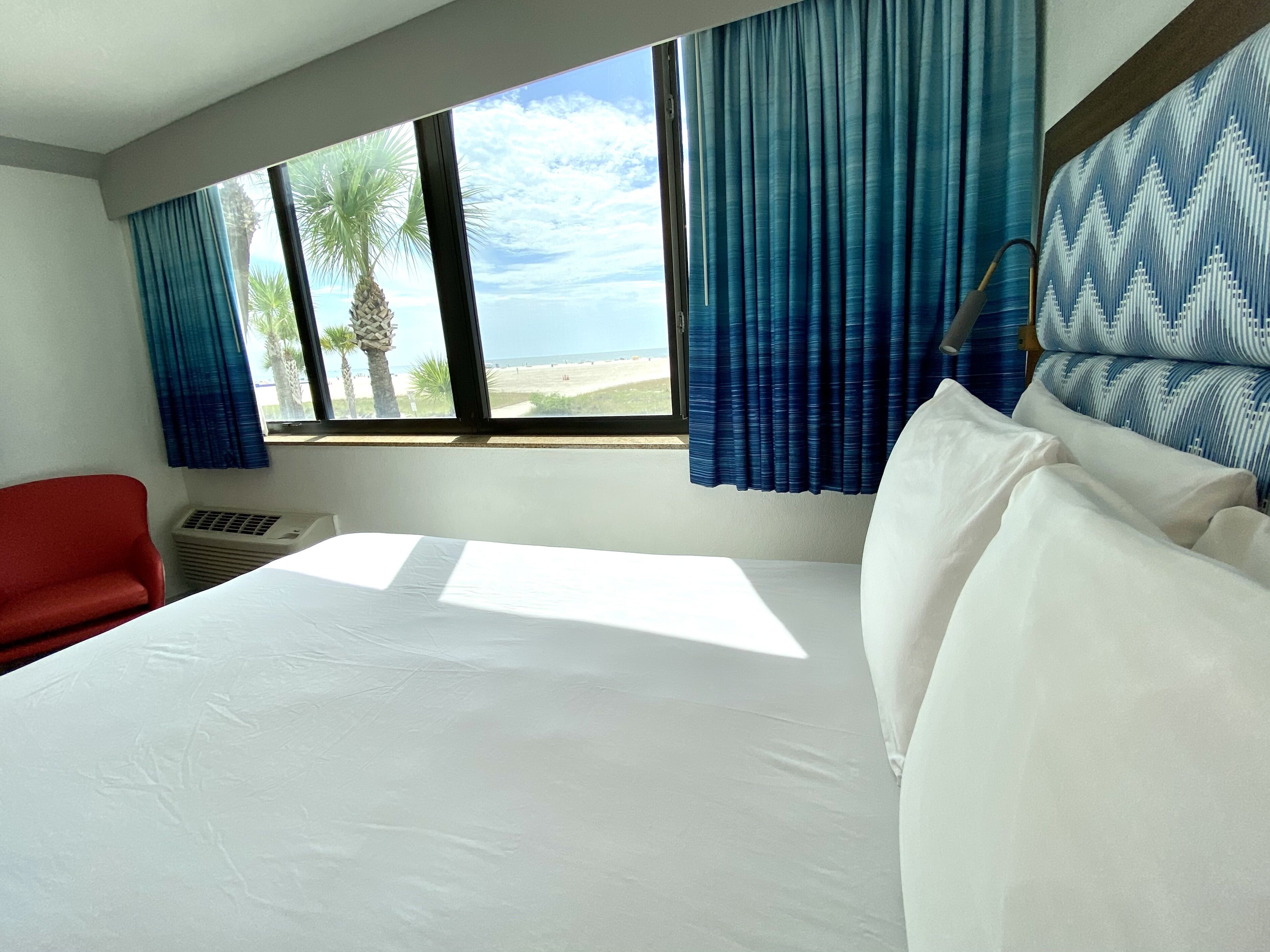 Une chambre d’hôtel avec un grand lit, un fauteuil rouge, une fenêtre donnant sur la plage et les palmiers, et des rideaux bleus.