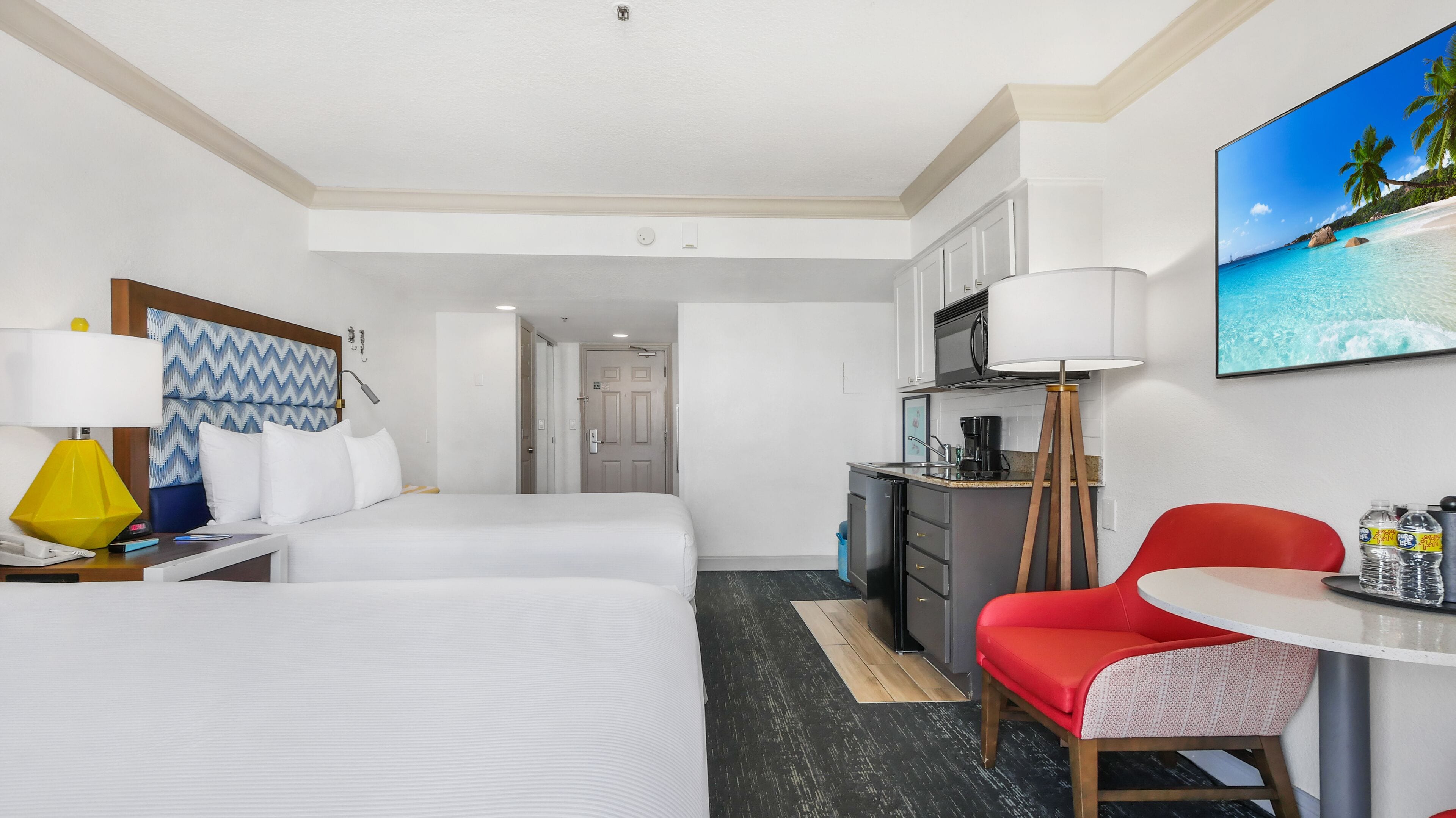 A hotel room with two beds, a desk with a lamp, a kitchenette, and a dining area with a red chair.
