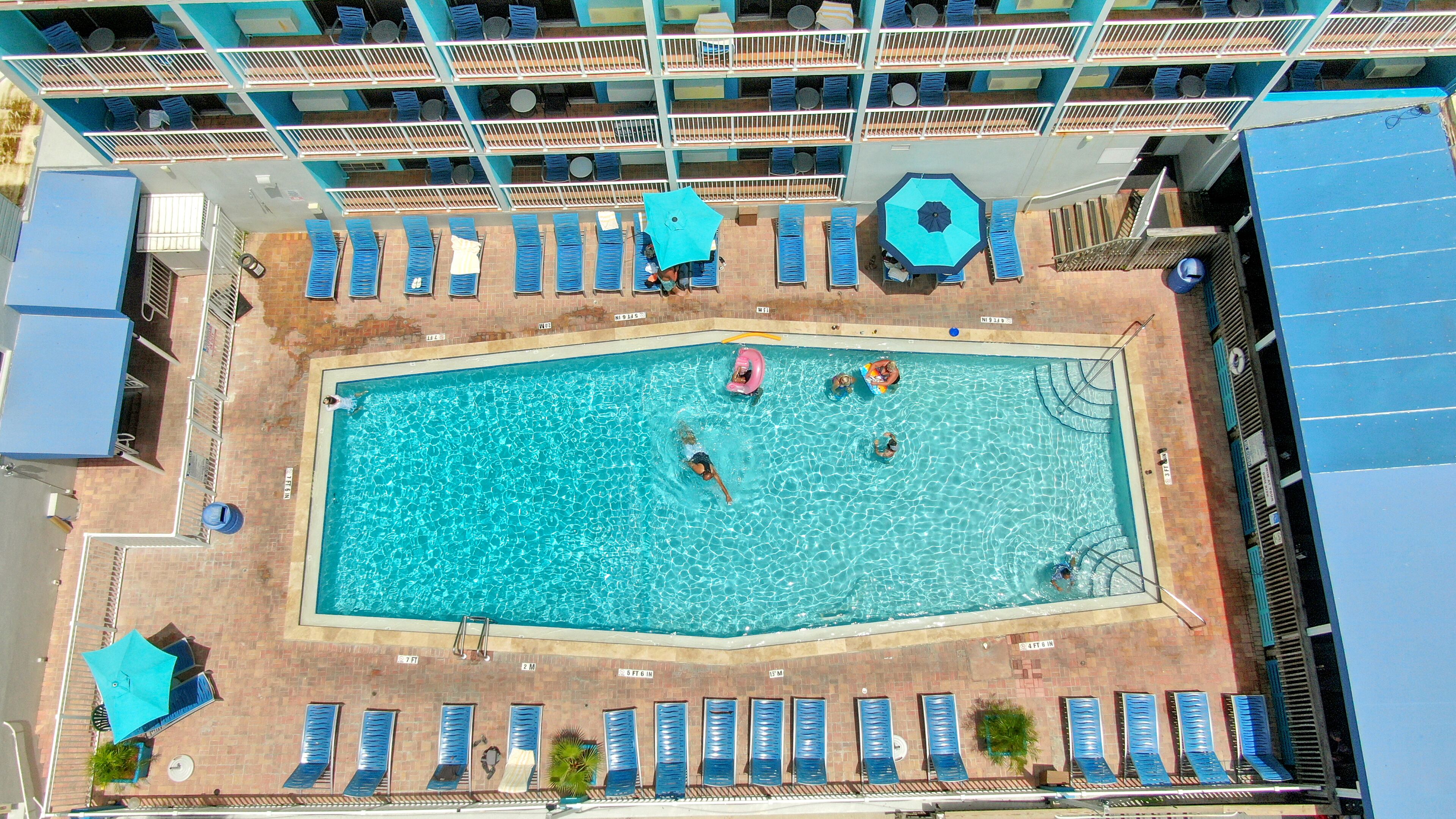 A rectangular swimming pool with a tiled deck, surrounded by blue lounge chairs and umbrellas.