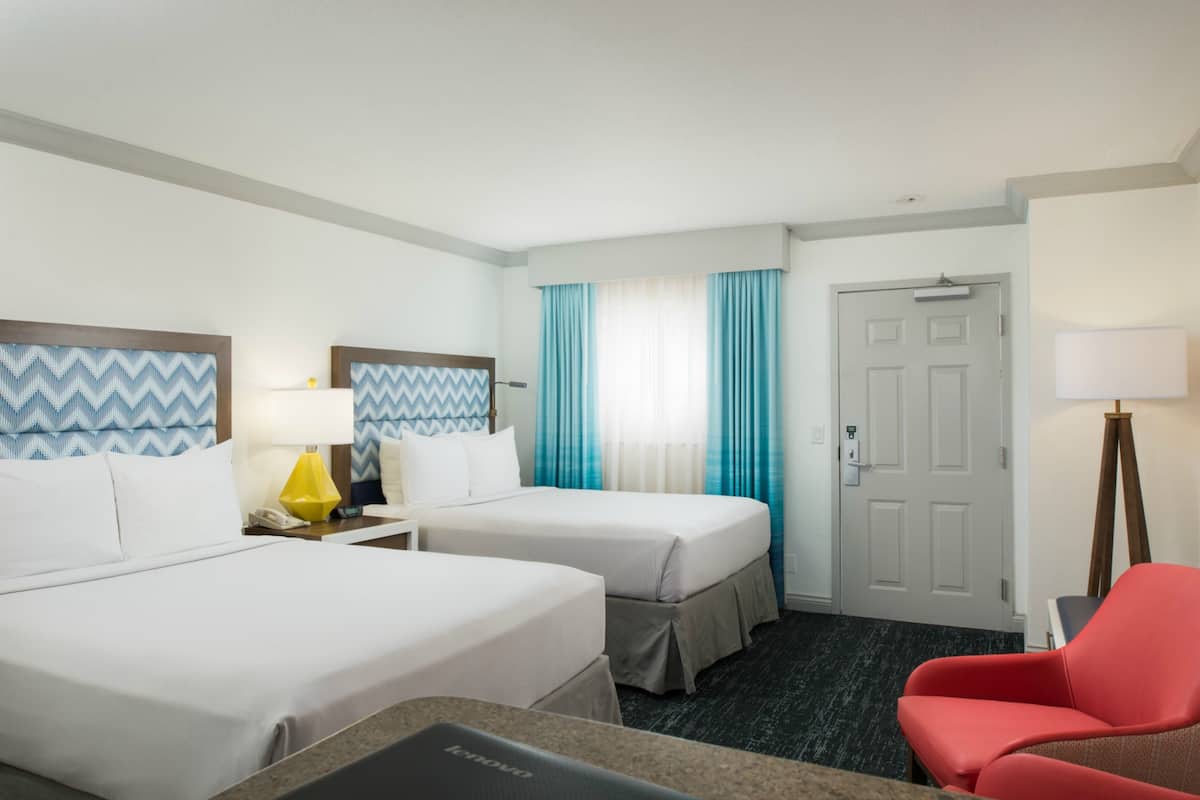 Quarto de hotel com duas camas, uma escrivaninha, uma cadeira e uma porta.
