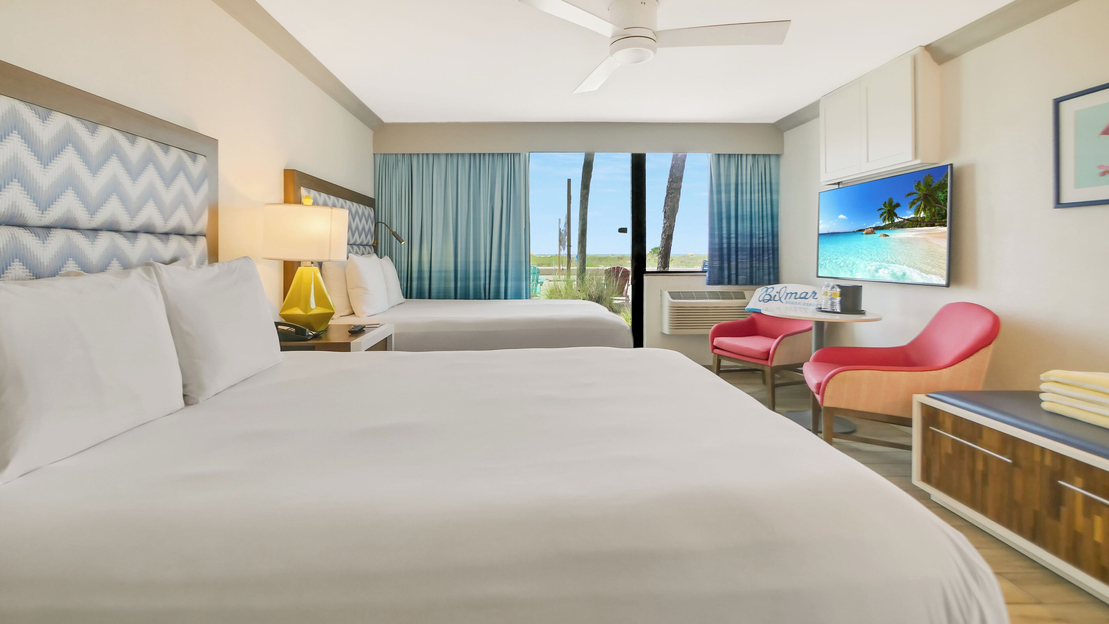 A hotel room with a bed, a desk with a chair, a TV, and a view of the beach.