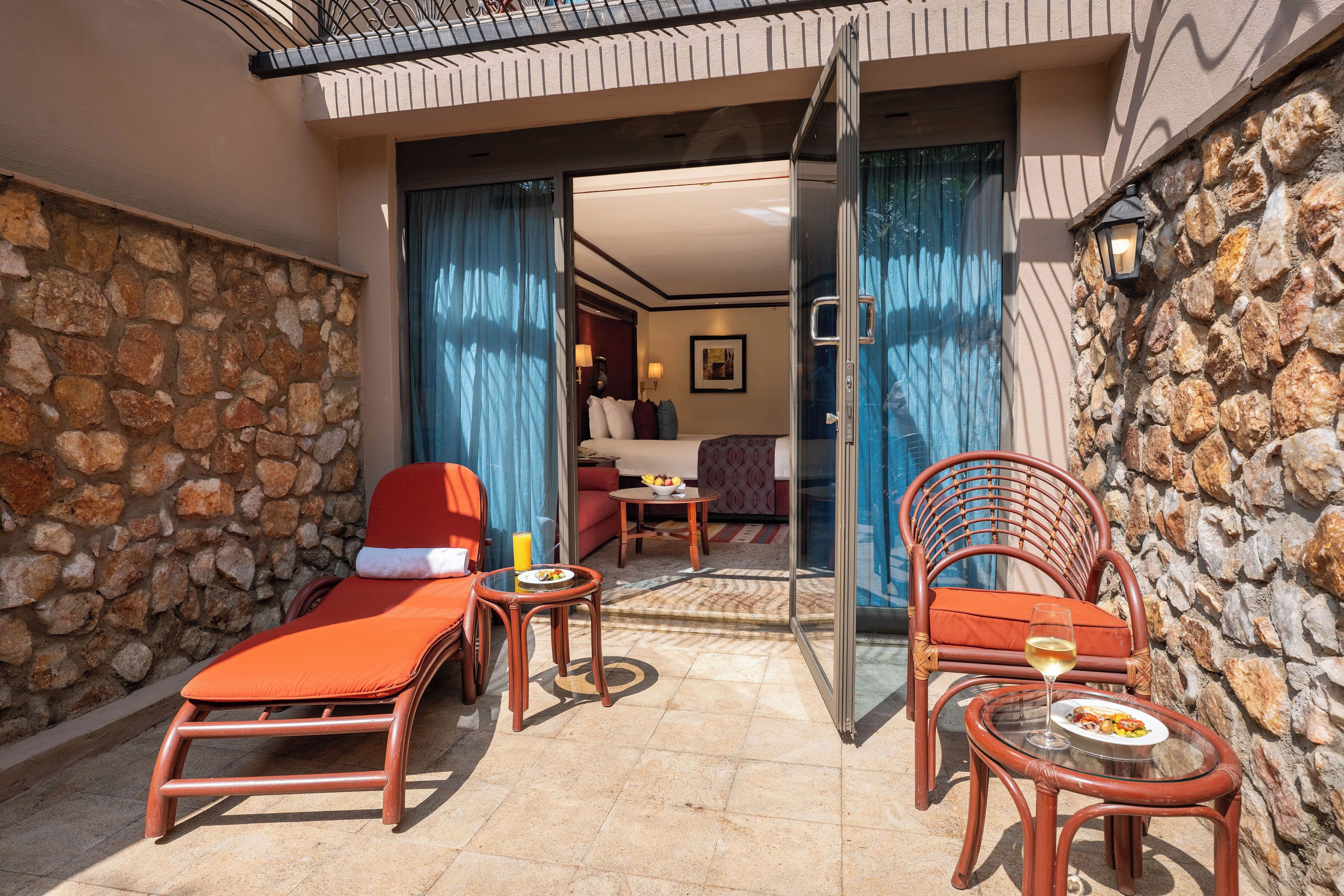 A sunlit outdoor seating area with lounge chairs, a table with a glass top, and a dining table with a plate of food and a glass of wine.