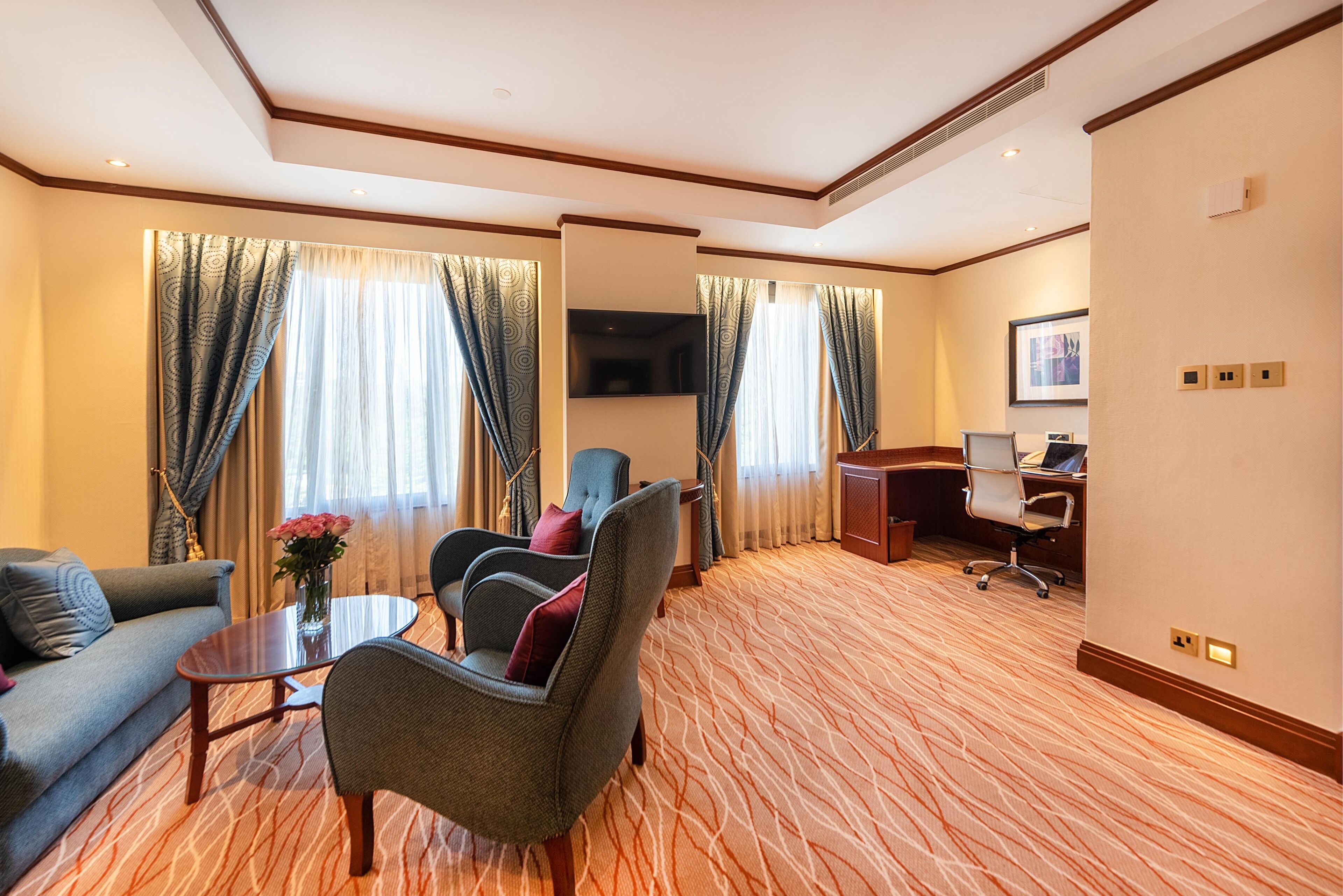A hotel room with a sofa, armchairs, a small table with a vase of flowers, a desk with a computer, and a large window with curtains.