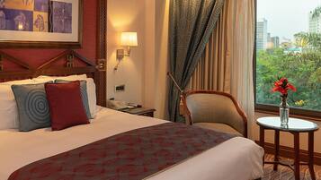 A hotel room with a large bed, a chair, a small table with a vase, and a window with a city view.