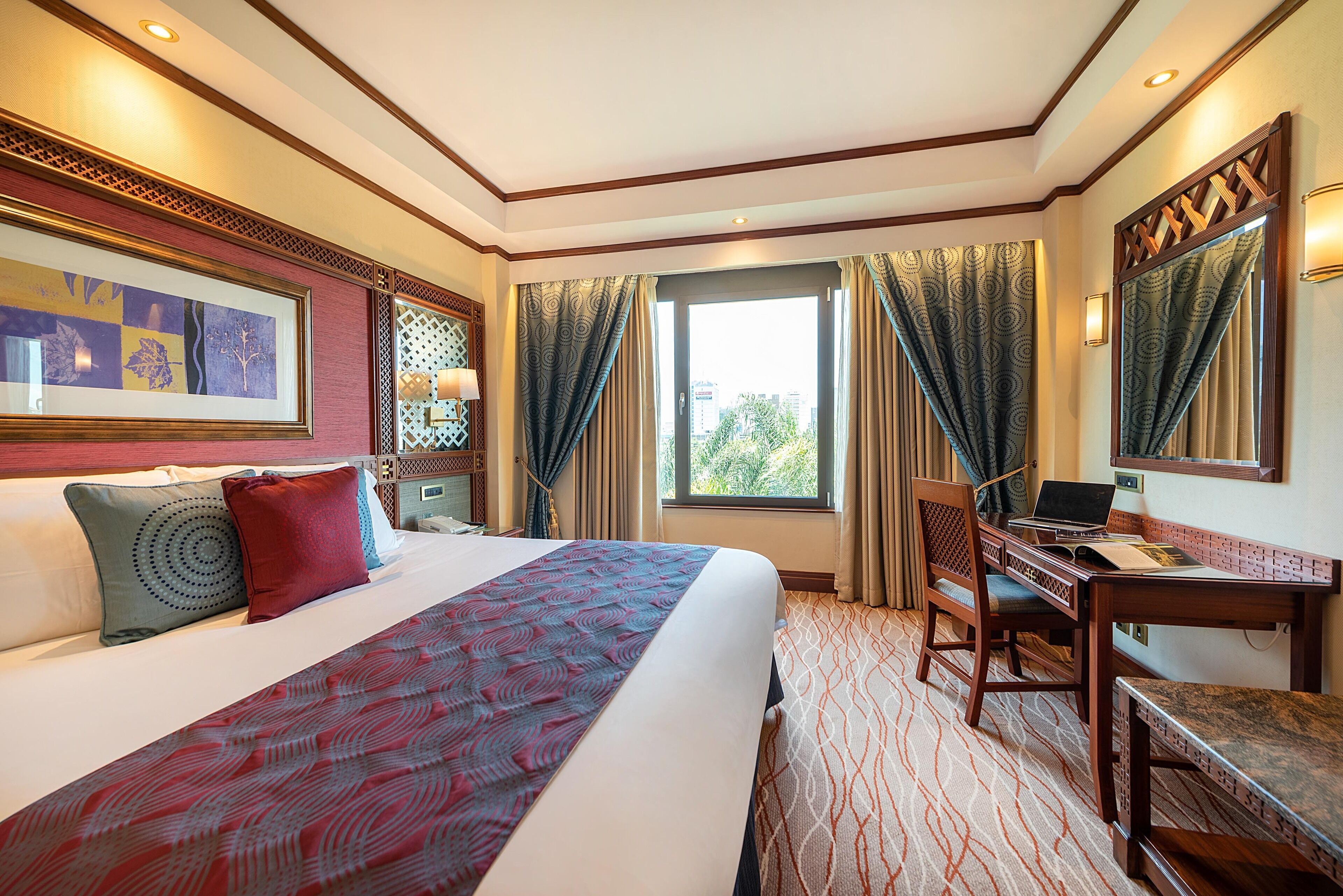 A hotel room with a large bed, a desk, and a chair. There is a window with curtains and a view of the outside.