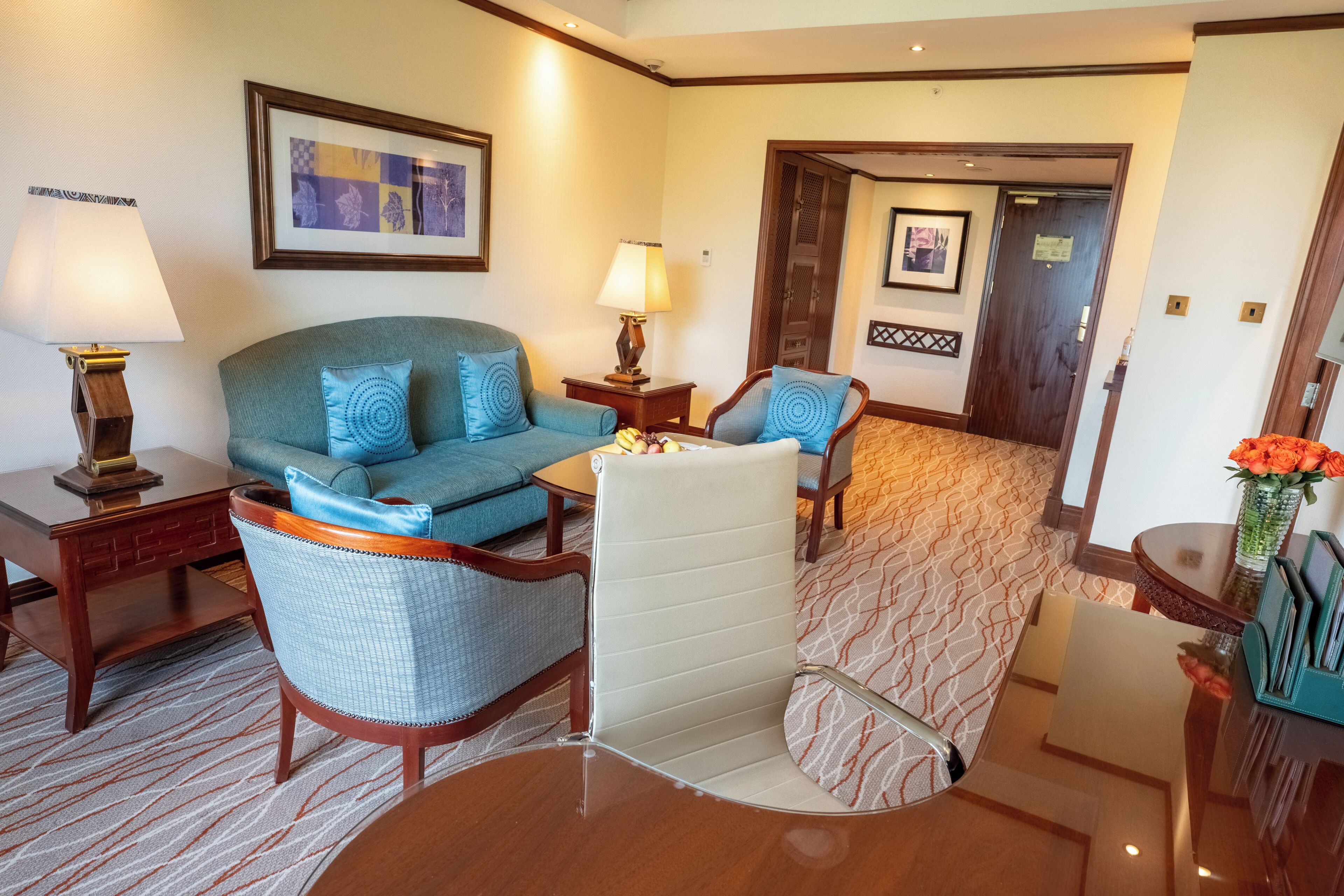 A hotel room with a sofa, armchairs, a small table, a lamp, and a framed picture on the wall.