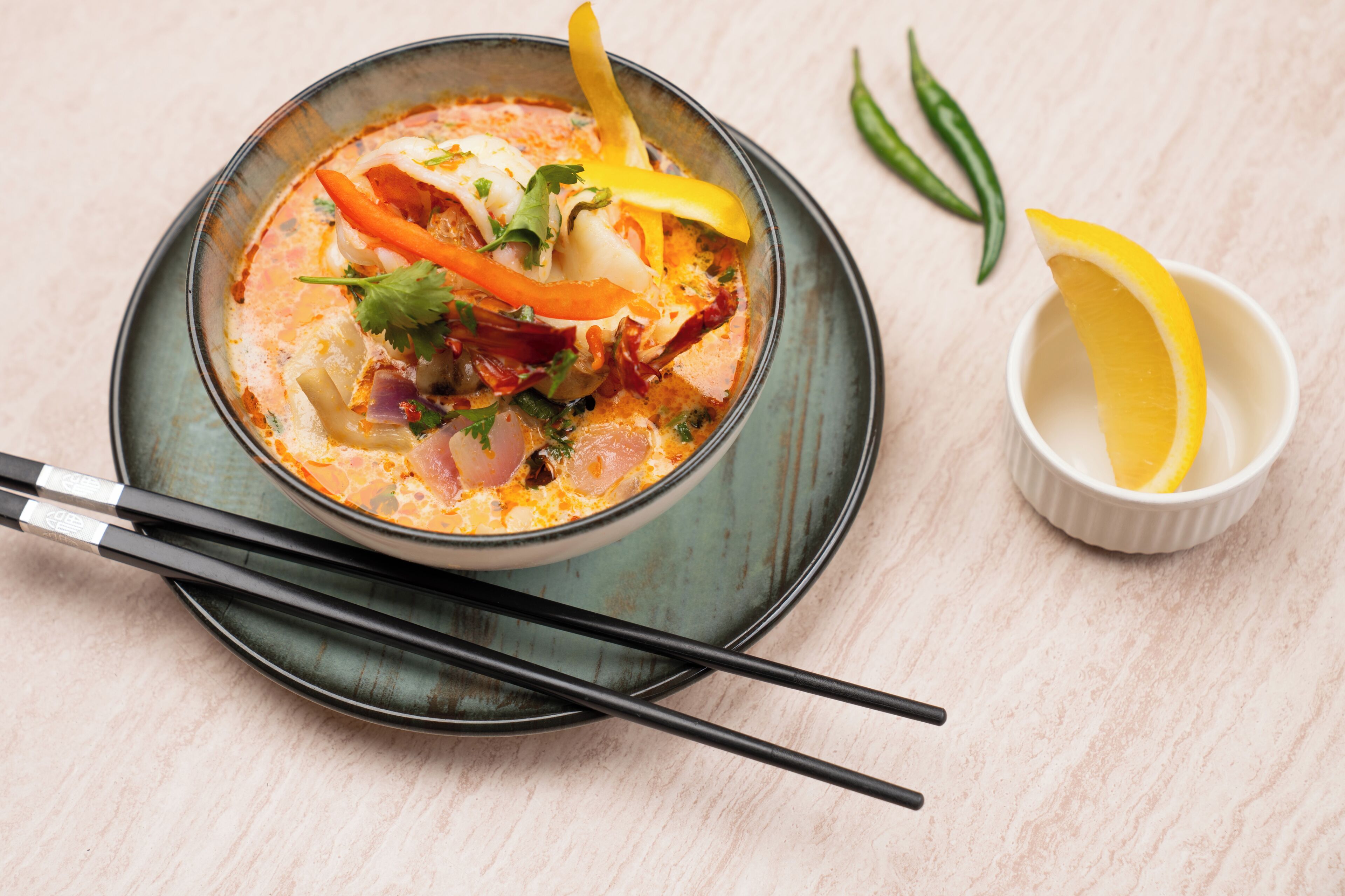 A bowl of soup with vegetables and herbs, a pair of chopsticks, and a small bowl with lemon slices.