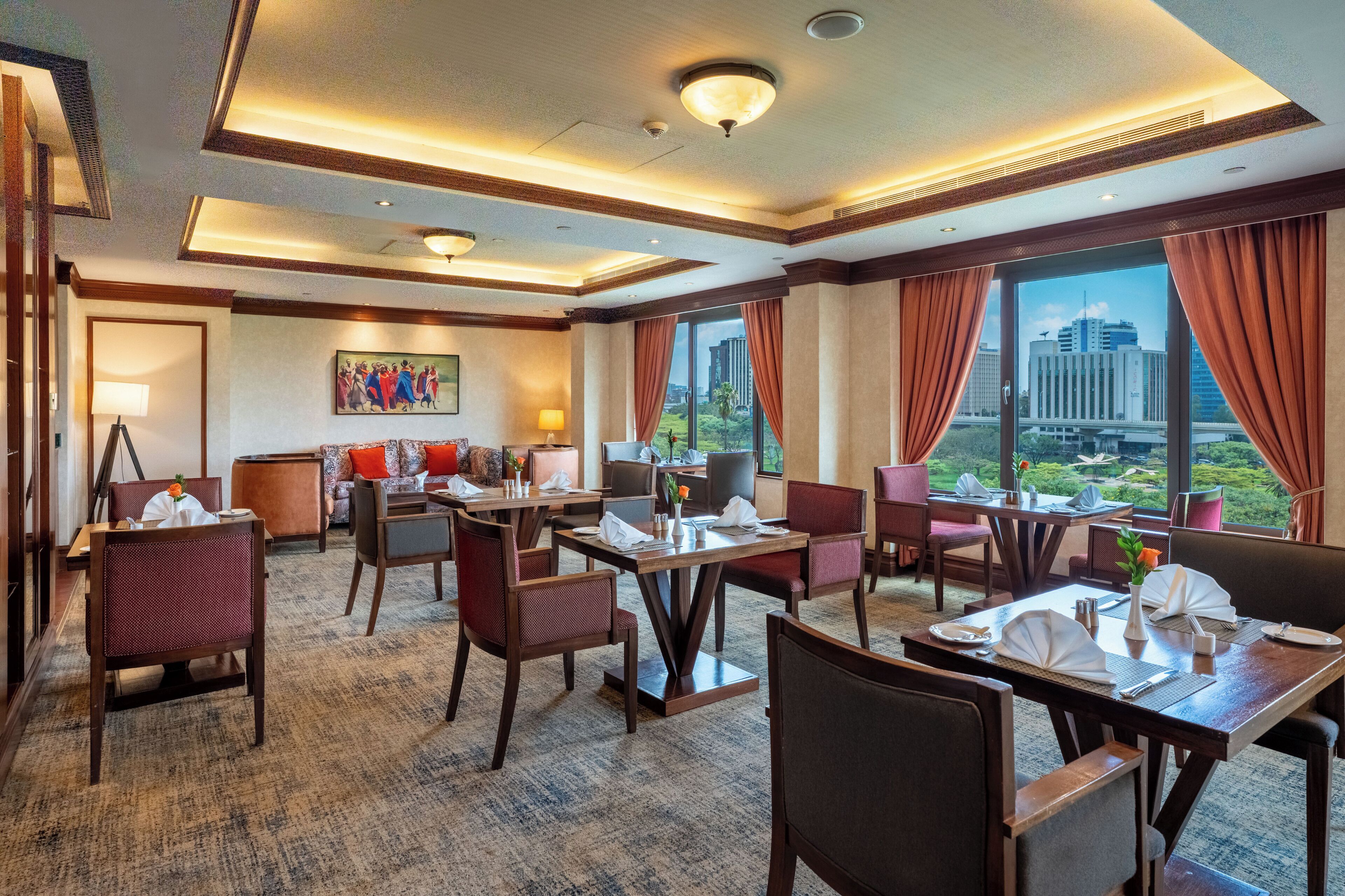 A hotel lobby with dining tables, chairs, a sofa, a painting, and large windows with curtains.