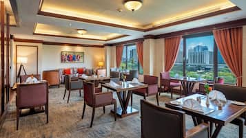 A hotel lobby with dining tables, chairs, a sofa, a painting, and large windows with curtains.