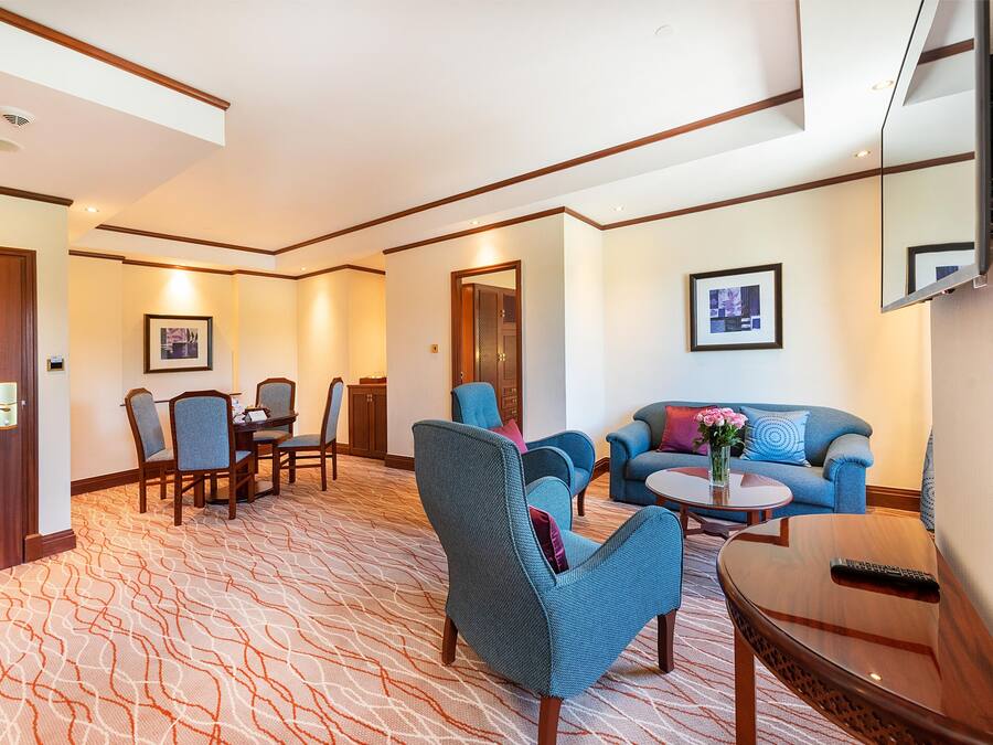 A hotel lobby with a patterned carpet, a dining area with a table and chairs, a sofa, and a TV mounted on the wall.