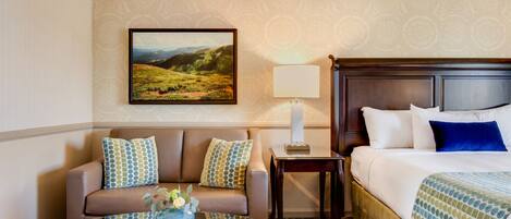 A hotel room with a sofa, a bed, a nightstand, a lamp, and a painting on the wall.