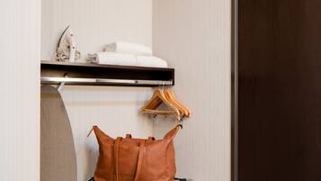 Habitación de hotel con una puerta de madera oscura, una pequeña zona de estar con un bolso de cuero marrón, un estante con toallas y un secador de pelo, y un perchero montado en la pared.