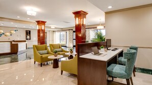 Un hall d’hôtel comprenant un comptoir d’accueil, deux fauteuils jaunes, une cloison en bois, un bureau avec deux écrans et une petite plante.