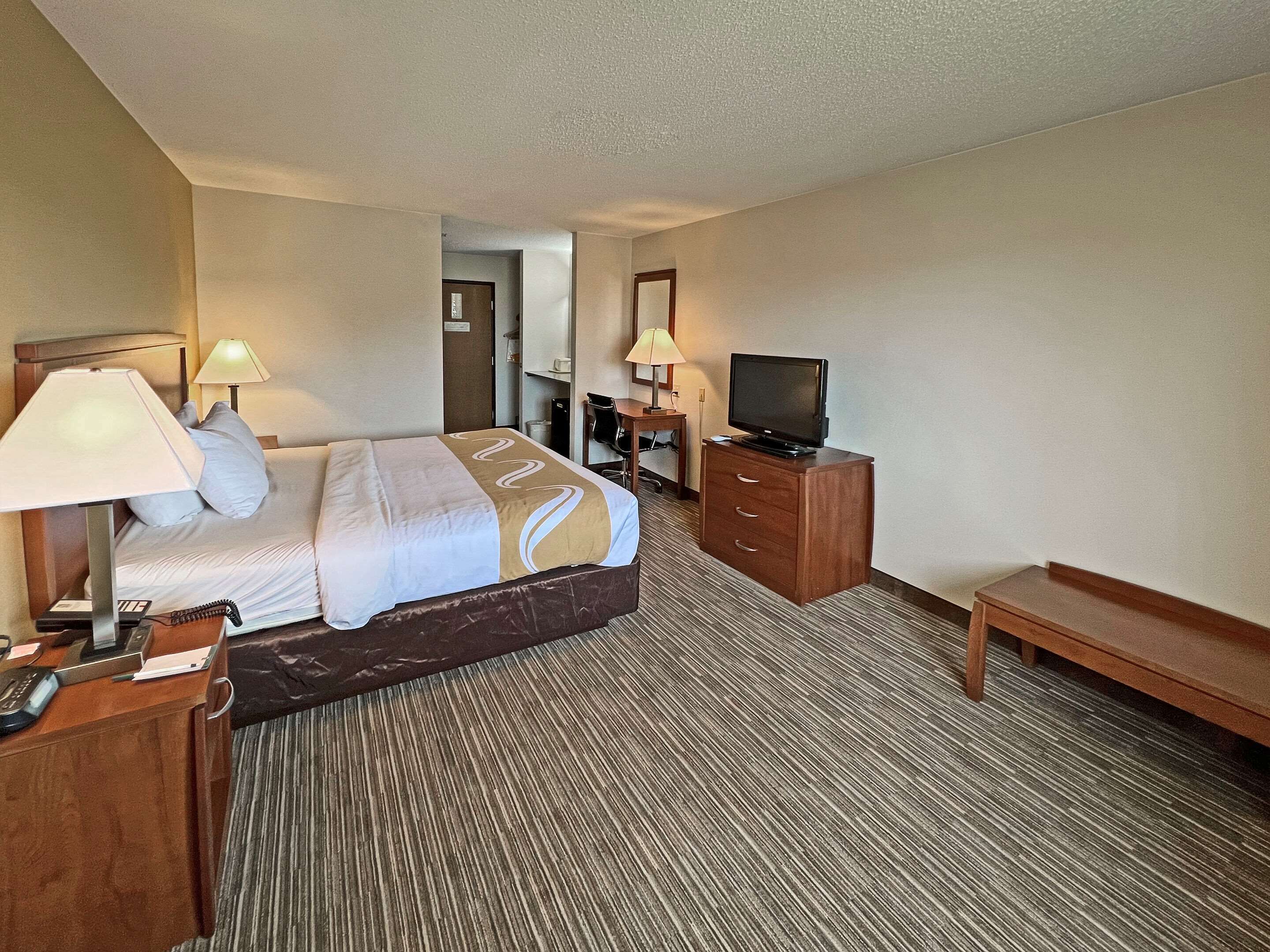 A hotel room with a bed, a TV, a desk with a lamp, and a chair.