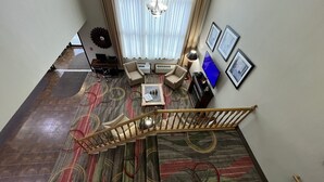 Un amplio salón con una escalera, un ventanal con cortinas, una araña y un televisor.
