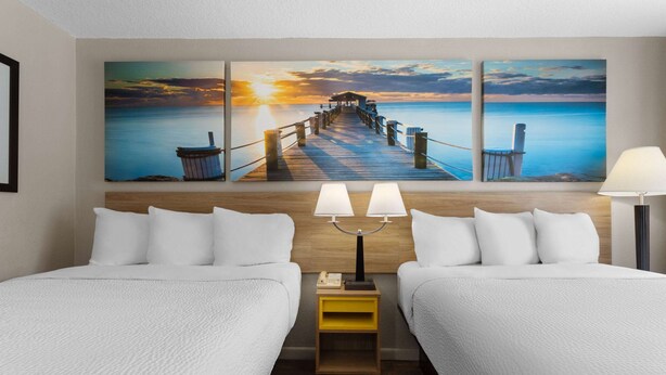Ein Hotelzimmer mit zwei Betten, einem Holzkopfteil, einem Nachttisch mit Lampen und einem Gemälde mit Sonnenuntergangblick an der Wand.