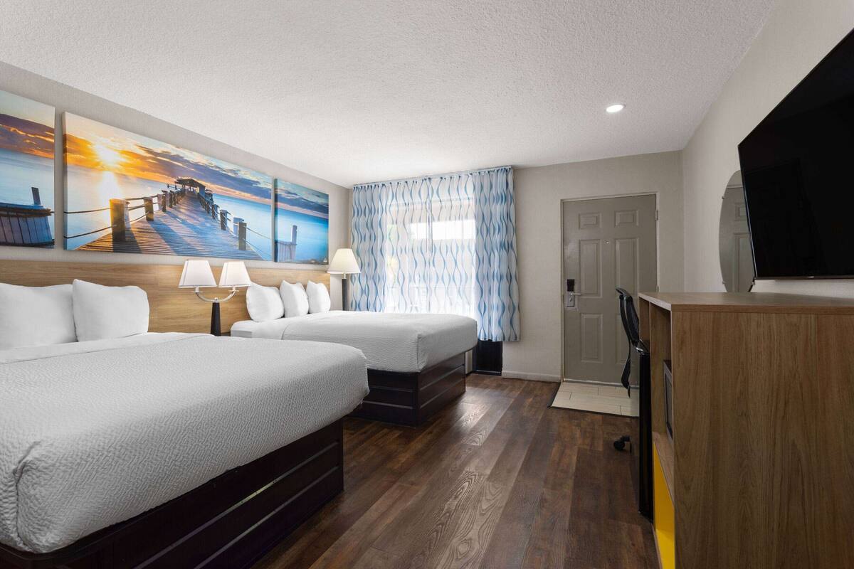 Ein Hotelzimmer mit zwei Betten, einem Holztisch, einem Flachbildfernseher und einem Gemälde, das einen Sonnenuntergang über einem Steg zeigt.