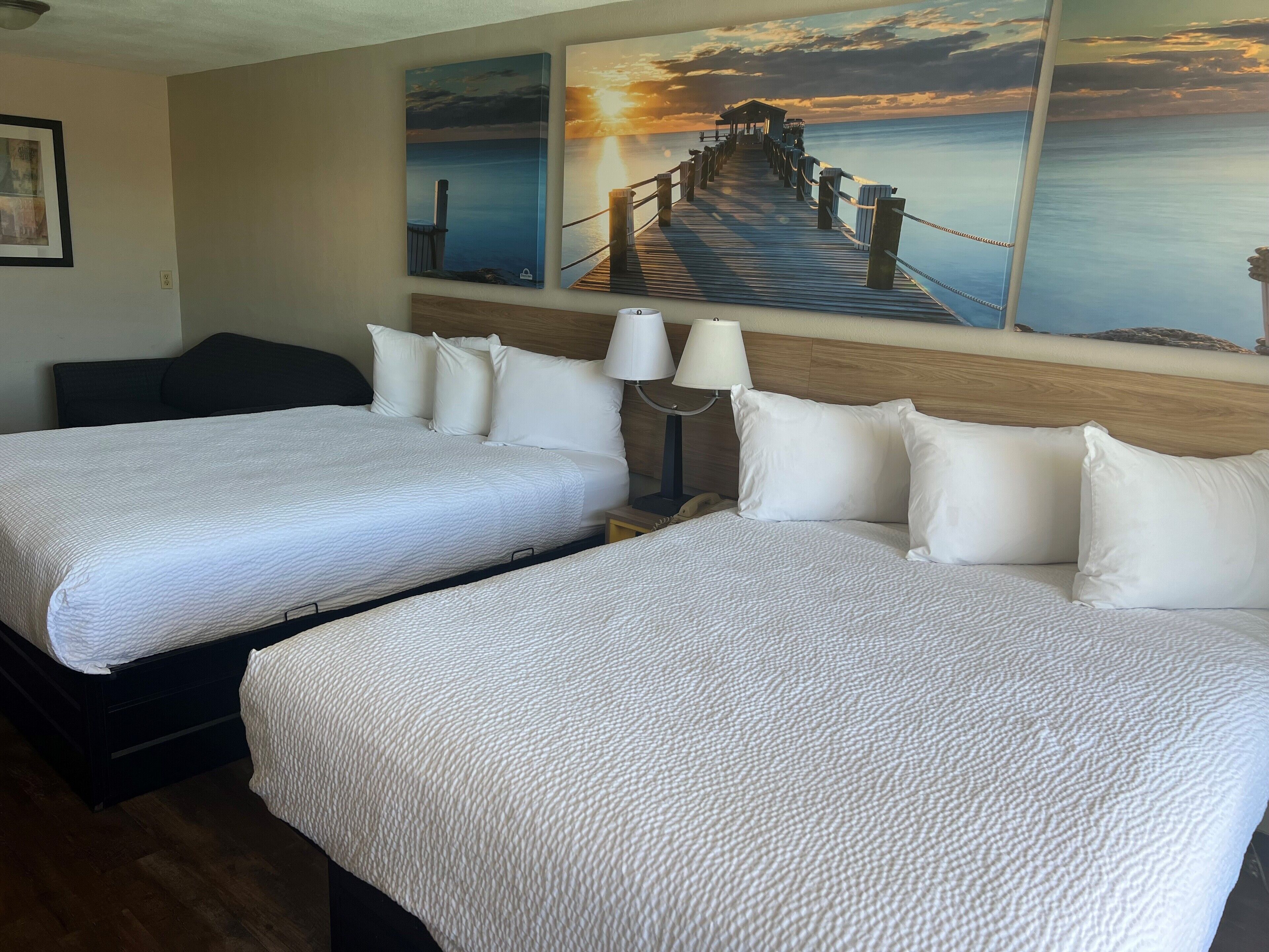 A hotel room with two beds, a nightstand with a lamp, and a painting of a pier at sunset.