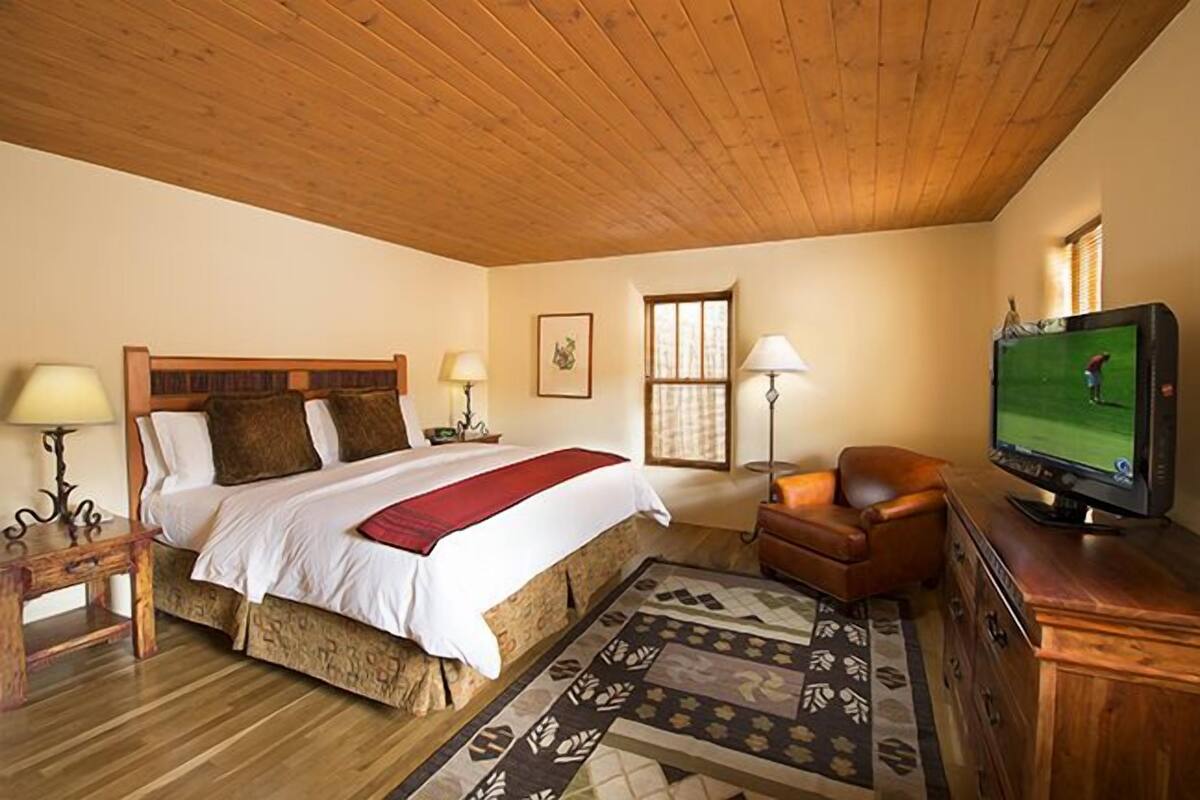 A bedroom with a wooden ceiling, a bed with a red blanket, a television, a chair, and a patterned rug.