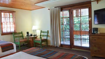 A hotel room with two beds, a wooden ceiling, a TV, a desk with a lamp, and a window with blinds.