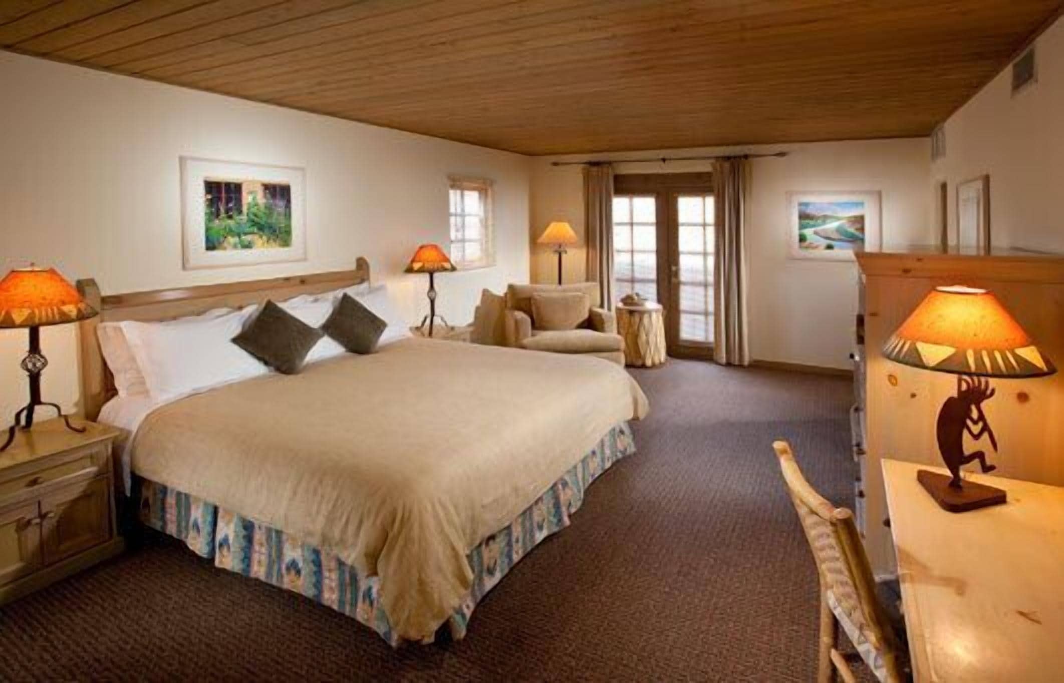 Une chambre d’hôtel avec un grand lit, deux tables de chevet avec des lampes, une chaise, une petite table et une fenêtre avec des rideaux.