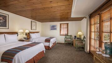 Quarto de hotel com duas camas, teto de madeira, obras de arte emolduradas, uma escrivaninha com abajur e uma janela com persianas.