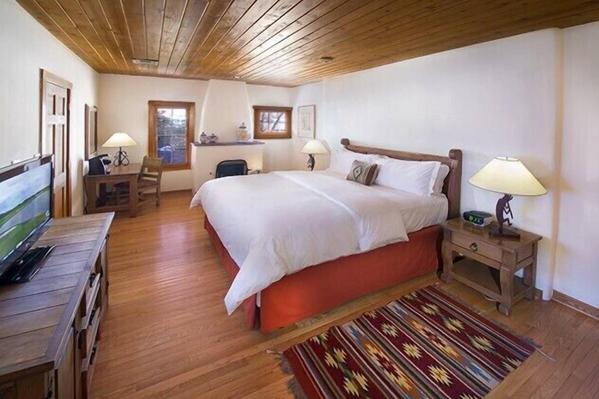 A bedroom with a wooden ceiling, a large bed with white bedding, two bedside tables with lamps, a desk with a chair, and a television on a wooden dresser.