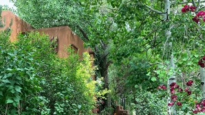 A stone pathway flanked by lush greenery and flowering plants, leading to a rustic building with a sloped roof. - Inn on the Alameda (Santa Fe)