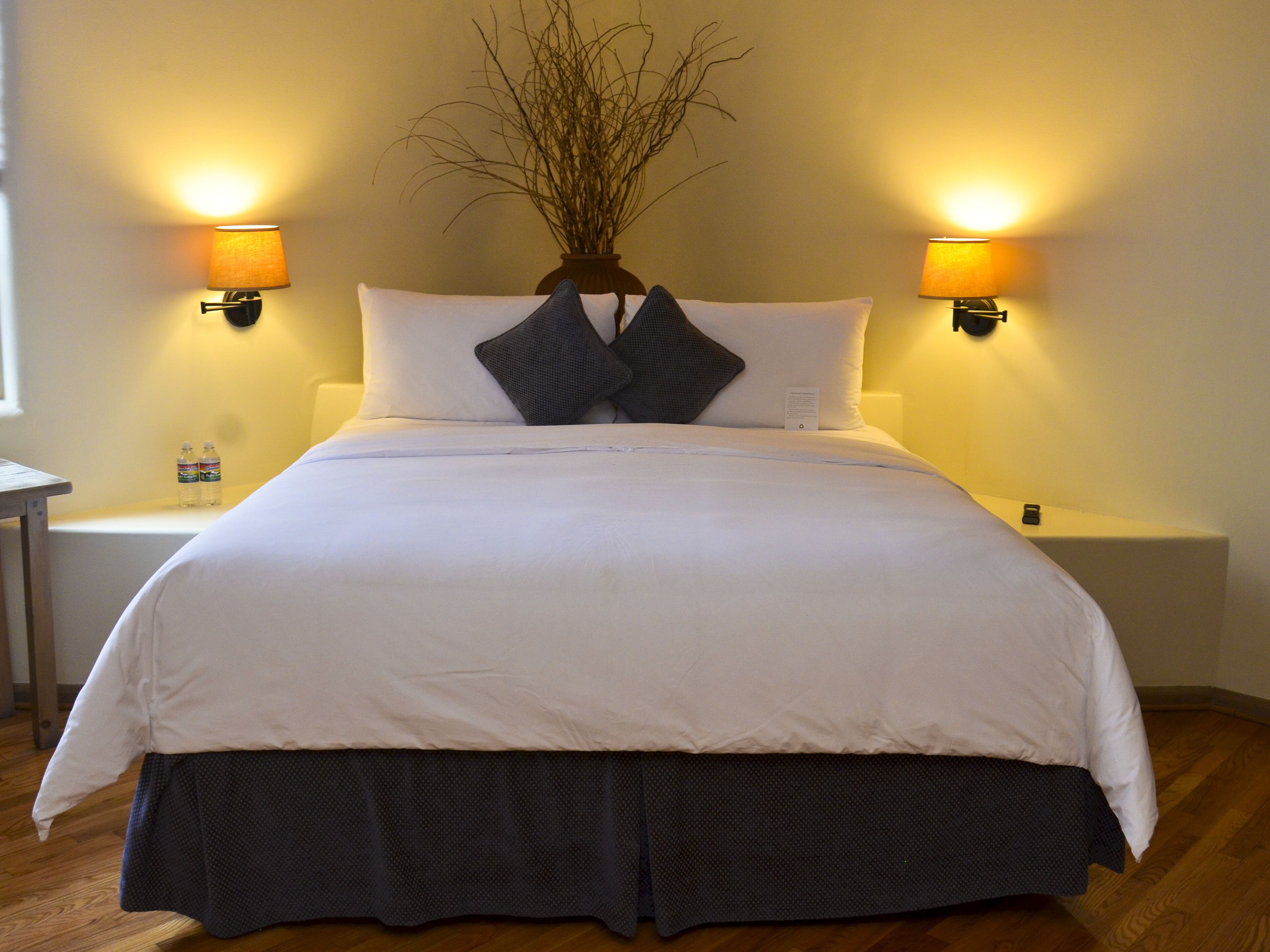 A neatly made bed with white and dark bedding, two wall-mounted lamps, and a vase with branches on the headboard.