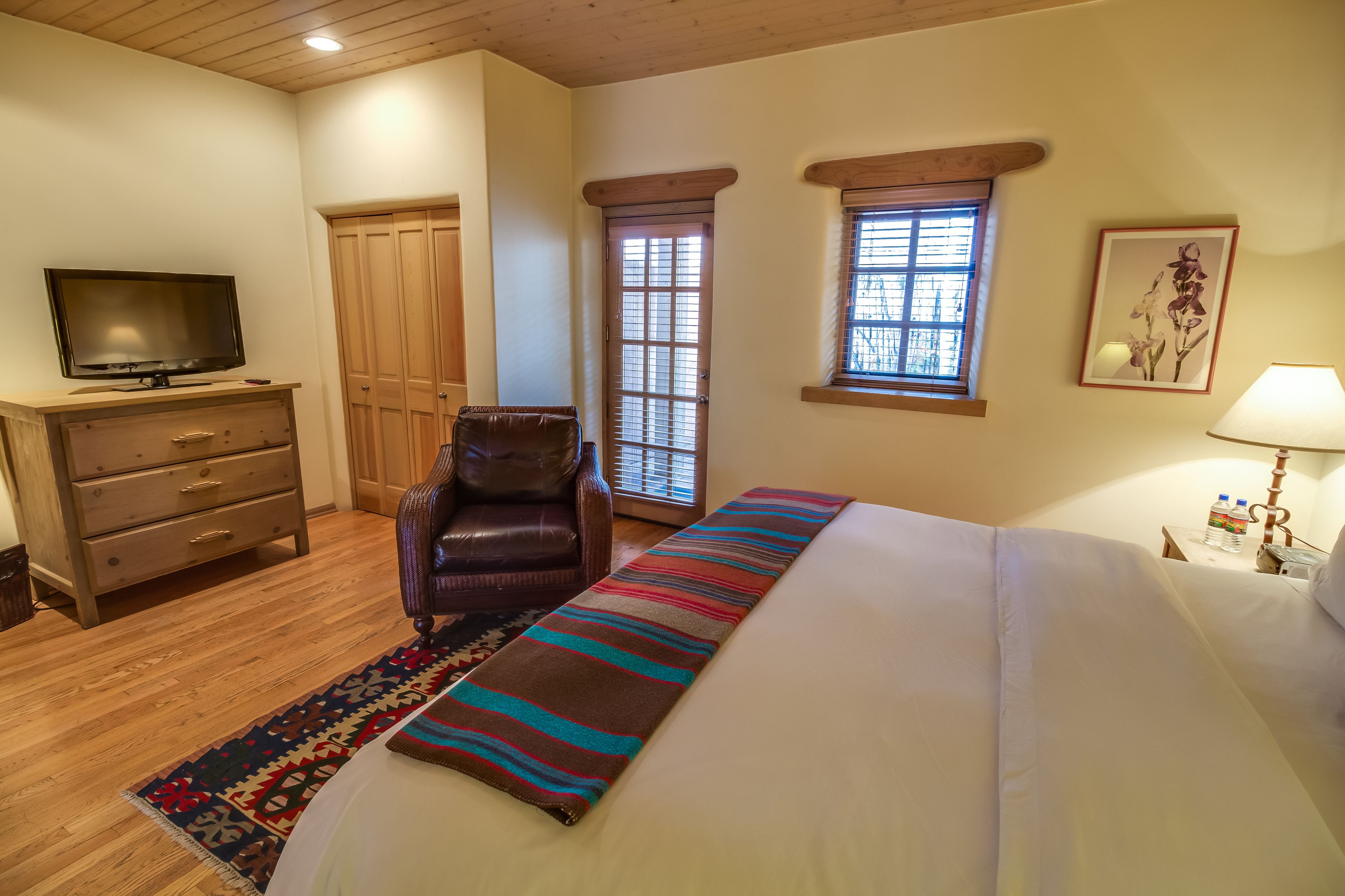 Un dormitorio con una cama, un televisor, un sillón de cuero, un aparador de madera, un tapete colorido, una ventana y un cuadro en la pared.