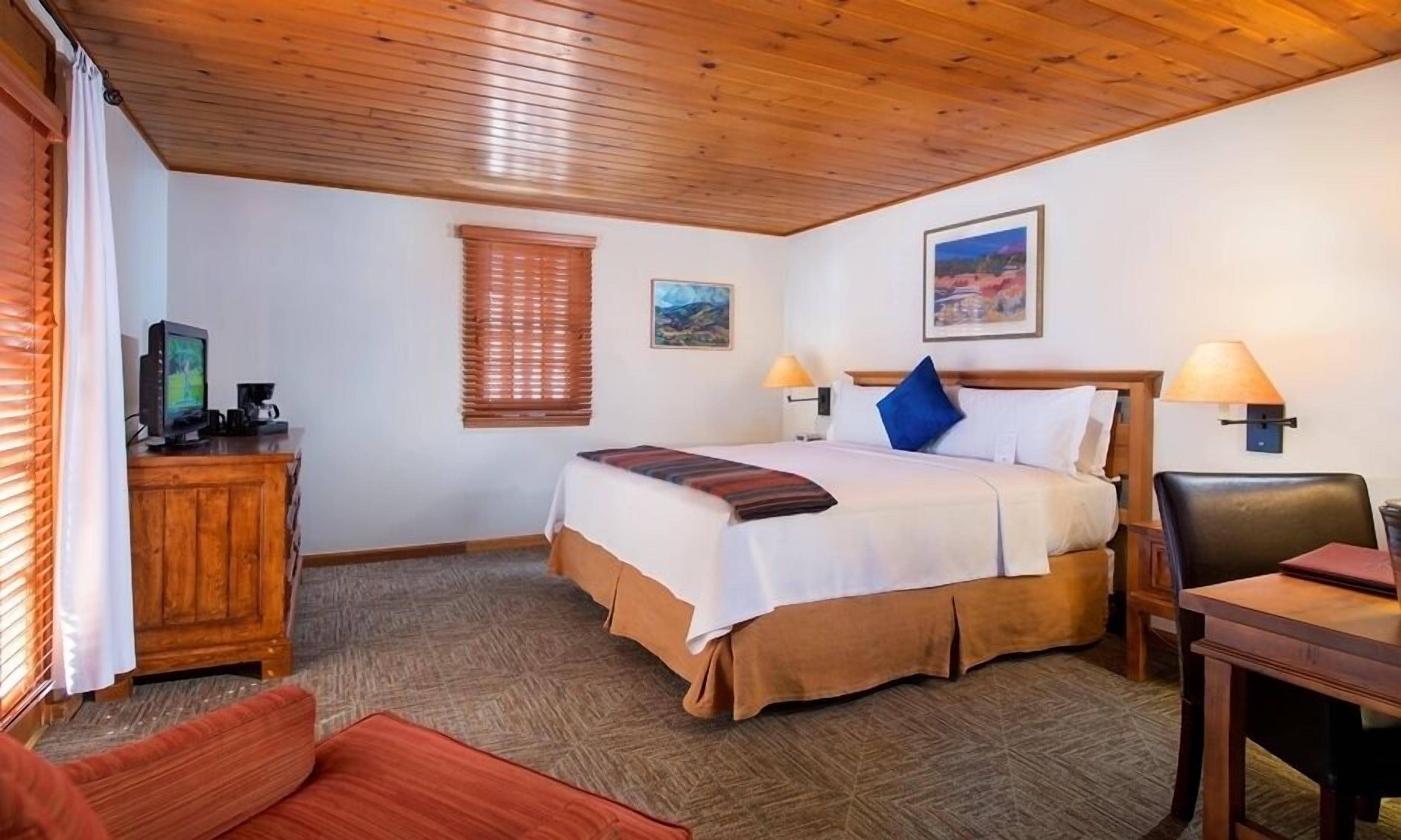 A hotel room with a wooden ceiling, a bed with a striped bedspread, two wall-mounted lamps, a desk with a chair, and a television on a wooden cabinet.