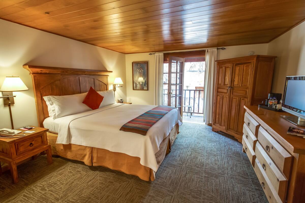 A hotel room with a wooden ceiling, a bed with white bedding and a red pillow, a nightstand with a lamp, a wooden dresser, and a television on the dresser.