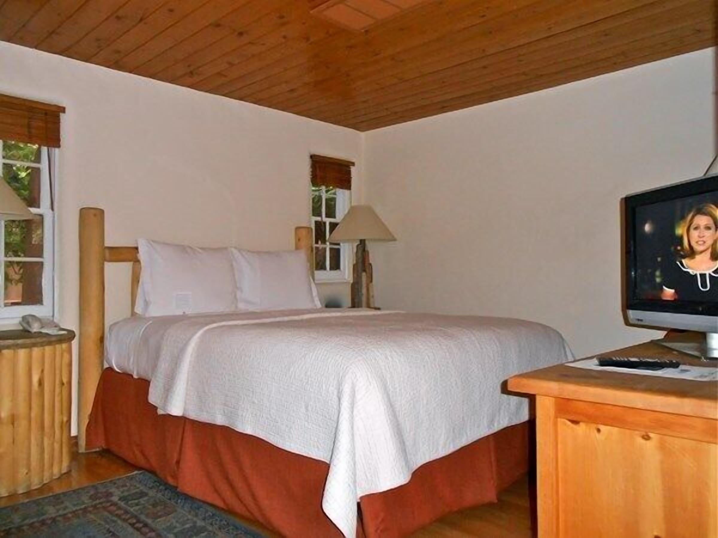 A bedroom with a bed, a television on a wooden table, and a window with blinds.