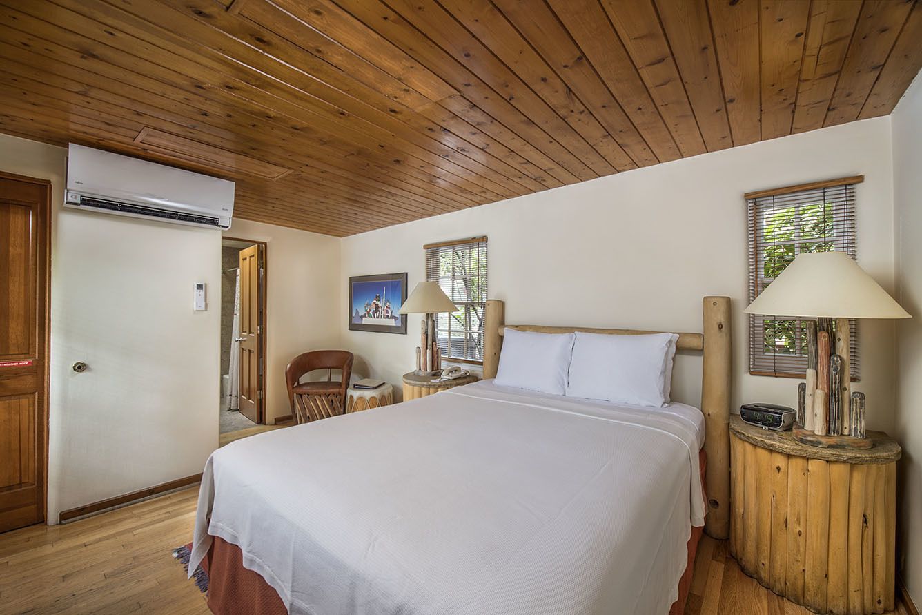 A bedroom with a wooden ceiling, a bed with white bedding, two bedside lamps, a window with blinds, and a door.