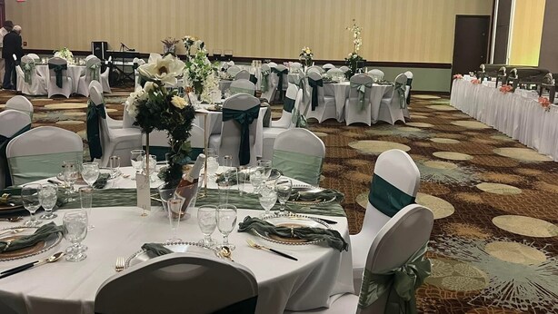 A banquet hall with round tables set for a formal event, featuring white tablecloths, green sashes, and floral centerpieces. - Lynchburg Grand Essential (Lynchburg)