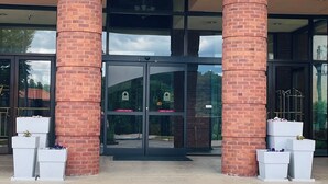 A building entrance with brick columns, glass doors, and white planters with flowers. - Lynchburg Grand Essential (Lynchburg)