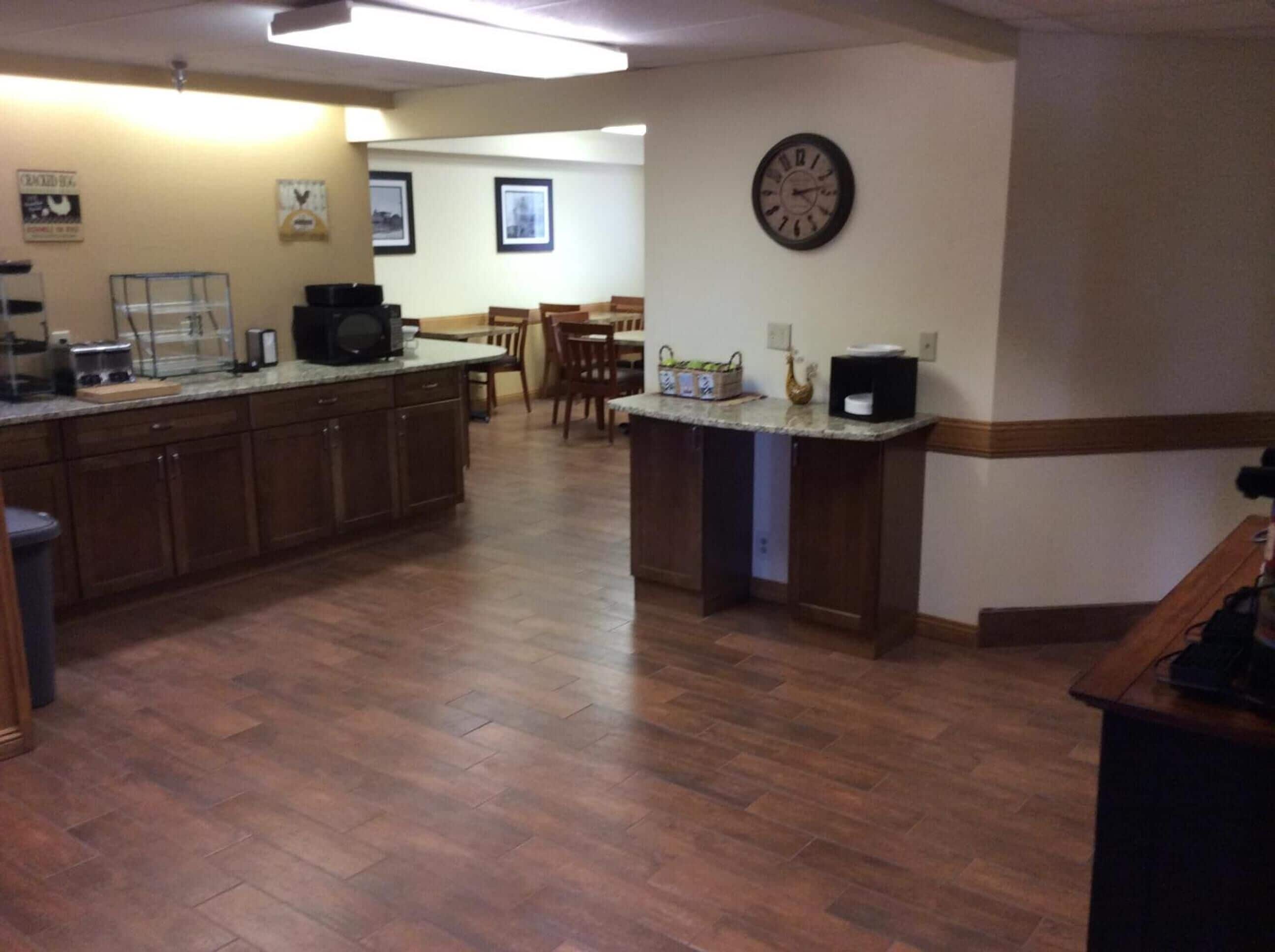 A kitchen with wooden cabinets, a microwave, and a coffee maker. There is a clock on the wall and a dining area in the background.