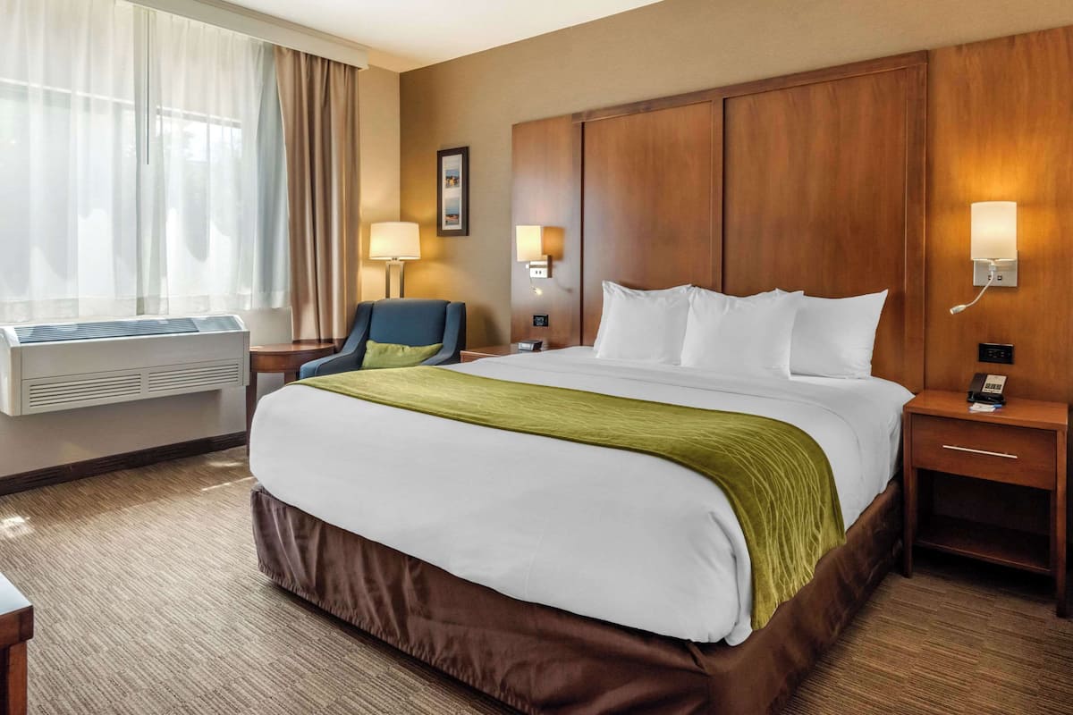 A hotel room with a large bed, a desk, a chair, a window with curtains, and two wall-mounted lamps.