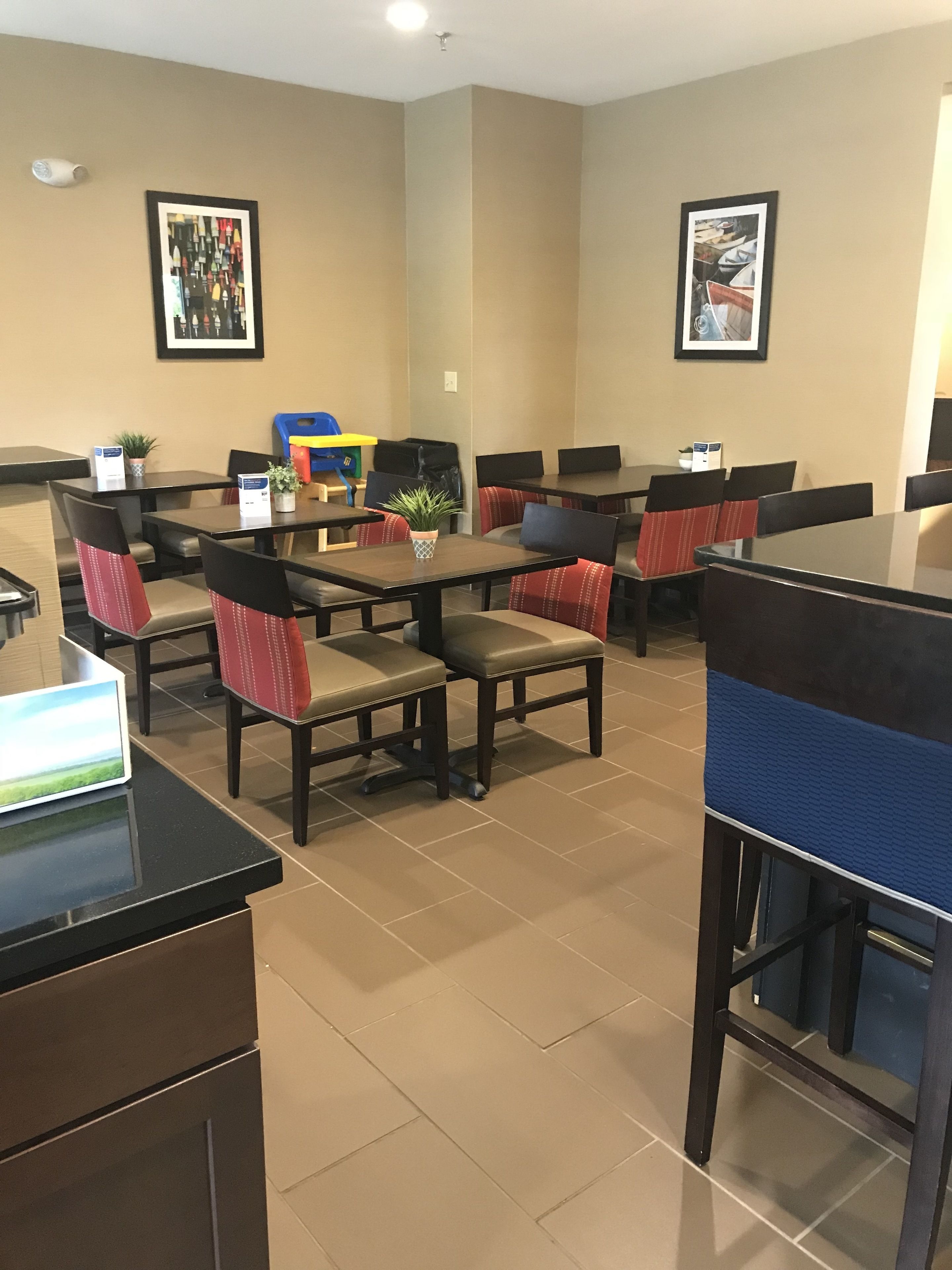 Une salle à manger d’hôtel avec plusieurs tables et chaises, dont certaines sont dotées de coussins rouges. Des tableaux encadrés ornent les murs et une petite plante est posée sur l’une des tables.