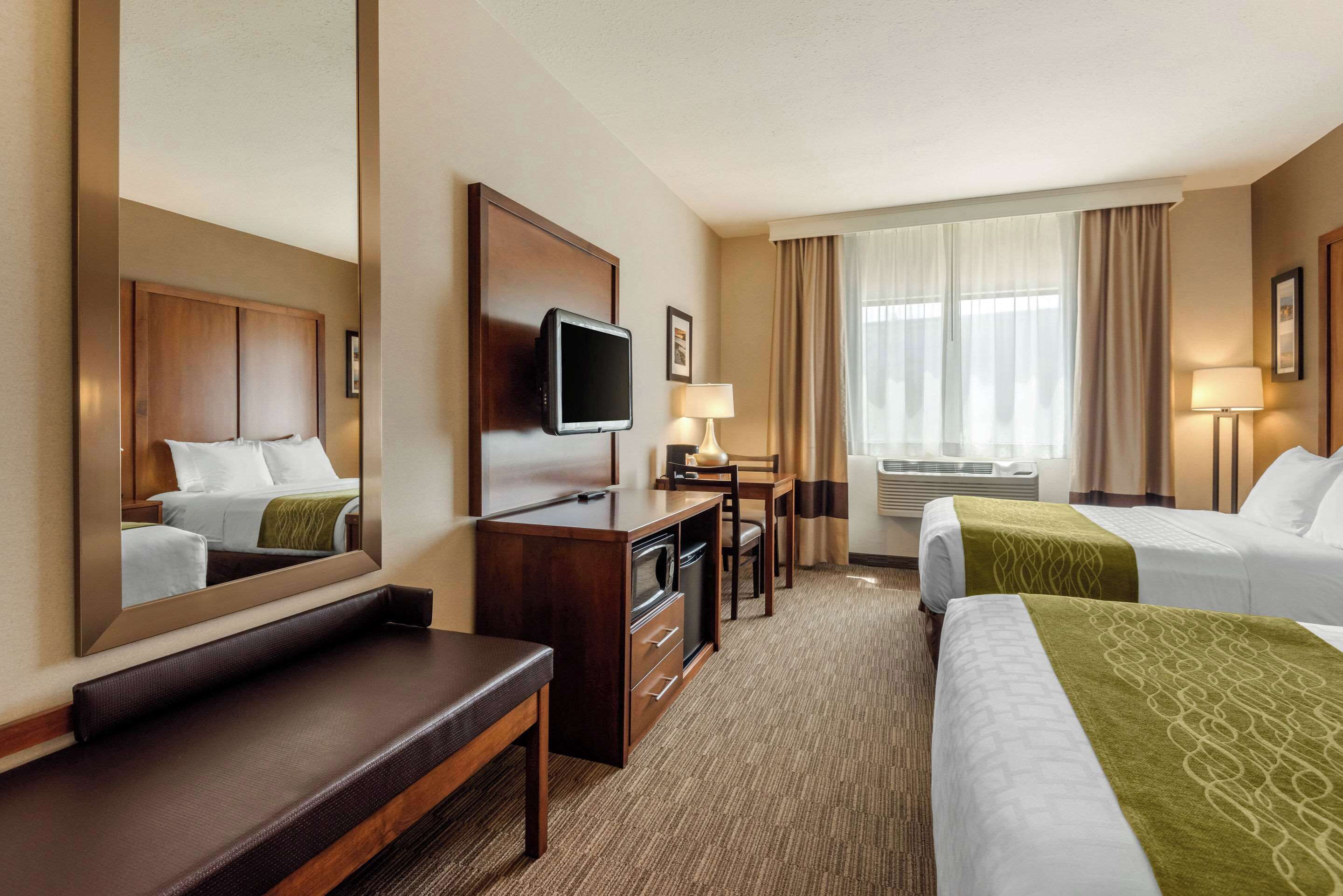 Ein Hotelzimmer mit einem großen Bett, einem Schreibtisch mit Fernseher, einem Spiegel, einem Fenster mit Vorhängen und einer Klimaanlage.
