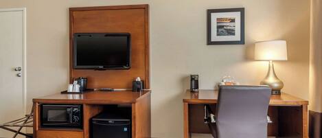 A hotel room with a wooden desk, a television, a coffee maker, a microwave, a mini fridge, a chair, and a lamp.
