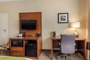 Habitación de hotel con escritorio de madera, televisor, cafetera, microondas, refrigerador pequeño, silla y lámpara.