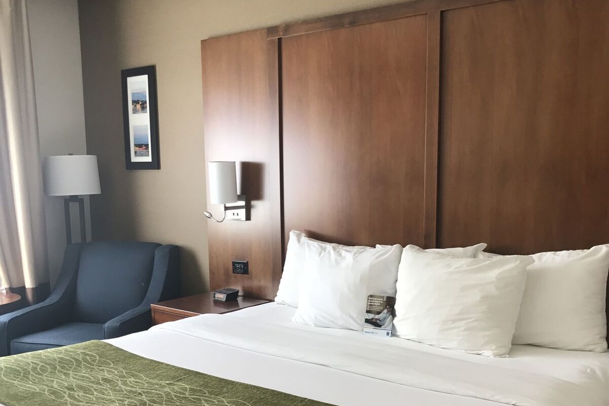 Una camera d'albergo con un letto grande, una sedia, un comodino, una lampada e un quadro appeso al muro.