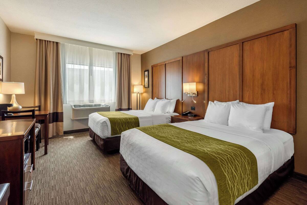 A hotel room with two beds, a desk, a chair, a lamp, and a window with curtains.