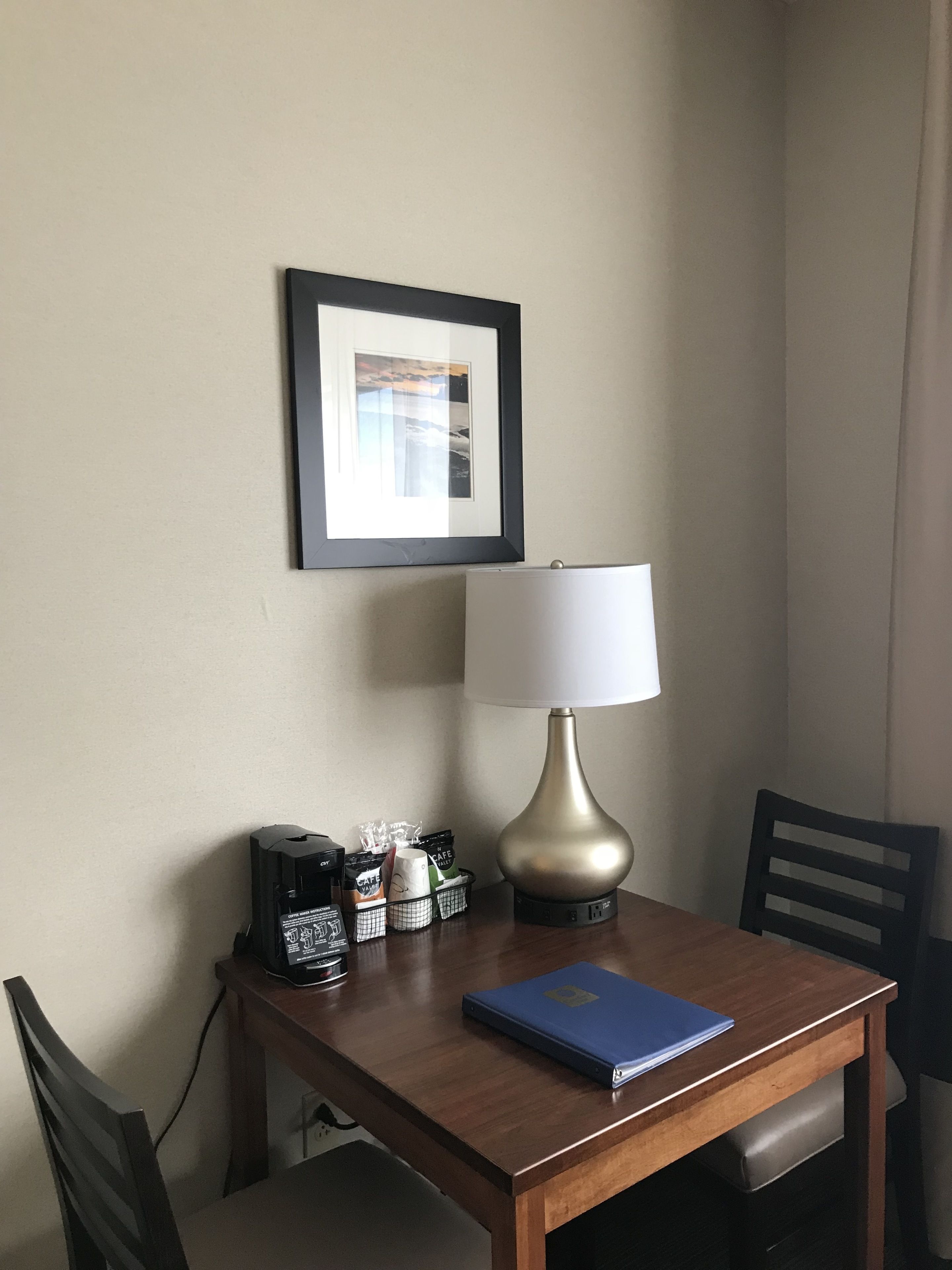 Une chambre d’hôtel avec une table en bois, une lampe, un tableau encadré et un dossier bleu.