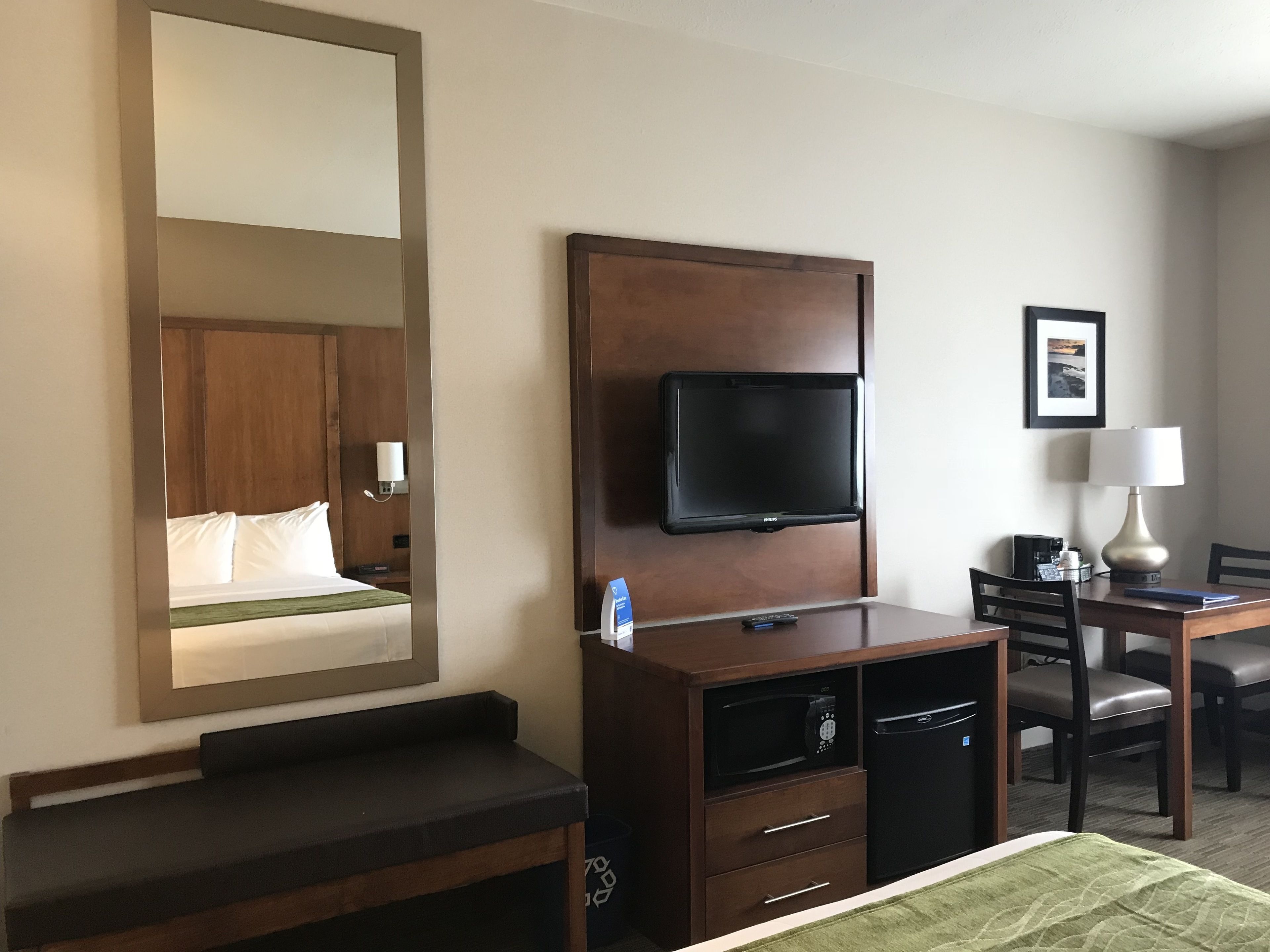 A hotel room with a bed, a TV mounted on a wooden panel, a desk with a lamp, and a chair.